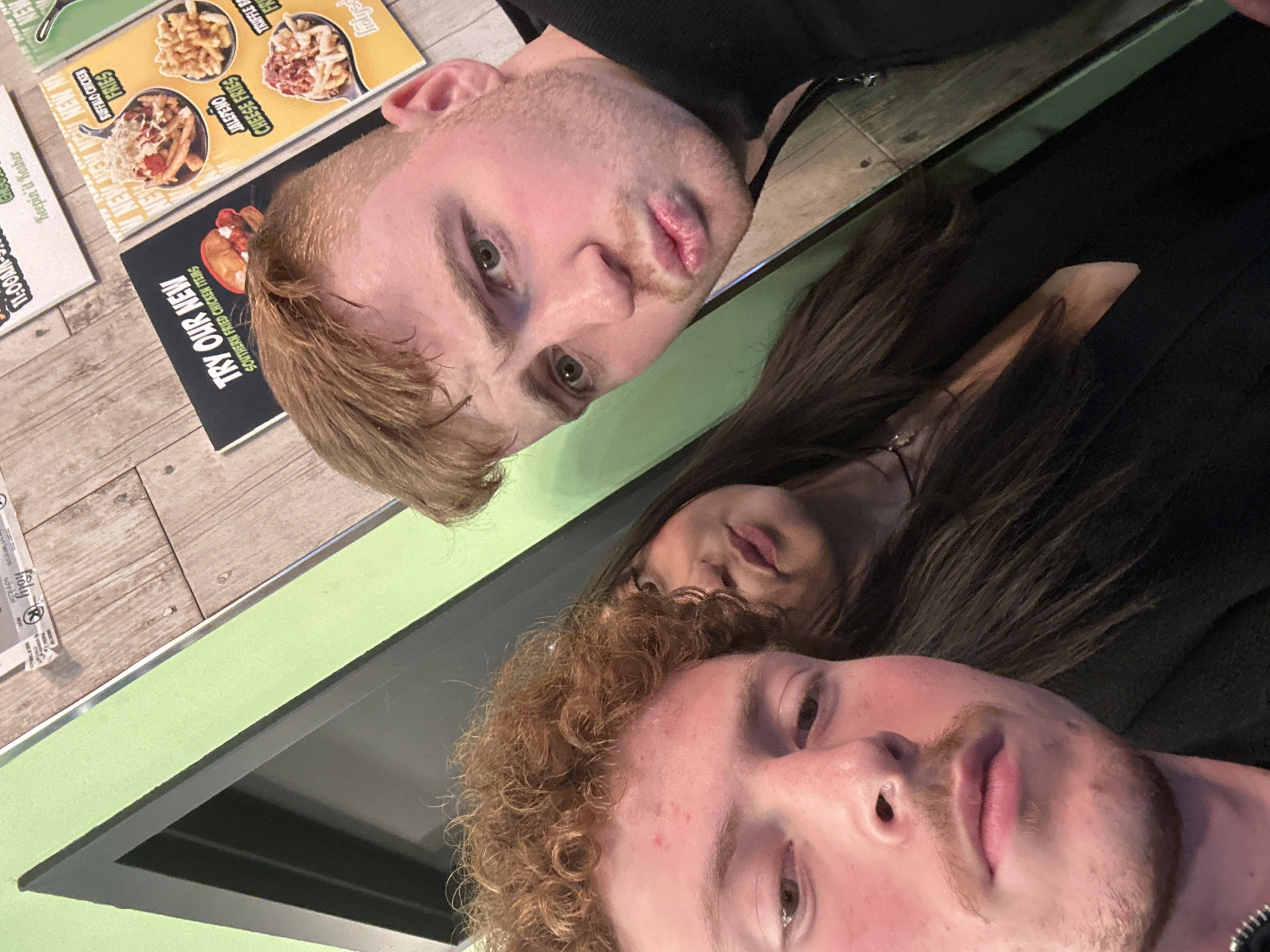 Three young adults are captured in a selfie-style photograph within a fast-casual restaurant setting. The location is identified as Holyschmoly, a Kosher establishment in New York, United States of America.
In the foreground left, a male with curly reddish-brown hair and a light mustache looks downwards, likely operating the camera. His body is largely obscured. To his right, partially visible, is a female with long straight dark brown hair, her head tilted, and lips slightly pursed towards the camera. In the foreground right, a male with short light brown hair and light facial stubble directs a "duck face" expression towards the camera. All individuals are wearing dark-colored clothing.
The background consists of two distinct wall sections. To the left, a vibrant lime green wall features a dark gray door frame. To the right, a wooden paneled wall displays multiple food-related posters. These posters advertise "TRY OUR NEW SOUTHERN FRIED CHICKEN ITEMS" with an image of a sandwich, and "NEW" loaded fries options including "BUFFALO CHICKEN FRIES," "JALAPENO CHEESE FRIES," and "TRUFFLE FRIES." Another poster specifies "Keepin' it Kosher" and lists operating hours, confirming the nature of the establishment.