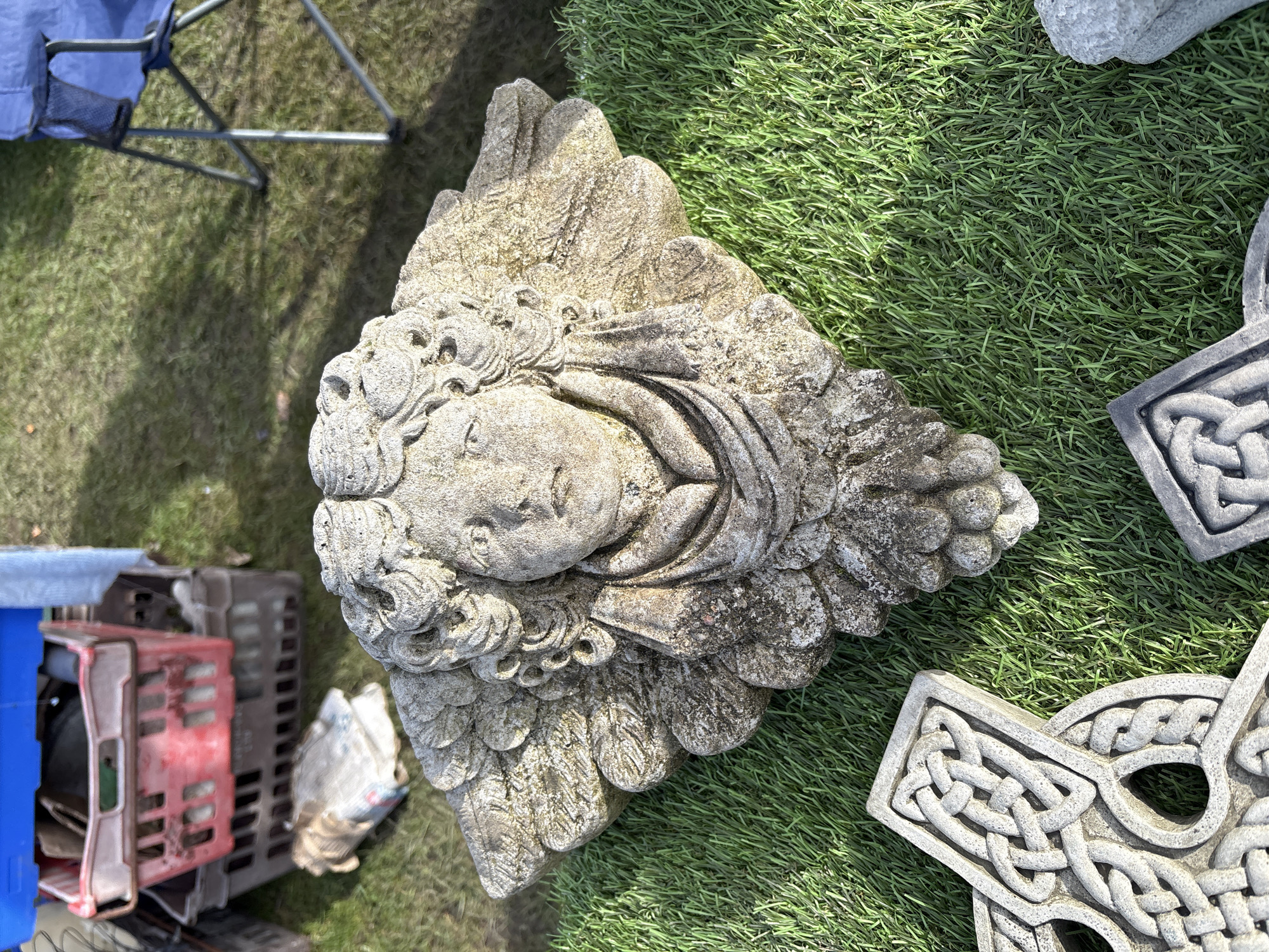 The image depicts a weathered, stone angel face sculpture lying on artificial green turf outdoors. Adjacent to the sculpture are stone Celtic crosses, also resting on the turf. In the background, there are crates and a folding chair. The location is Chirk, United Kingdom.