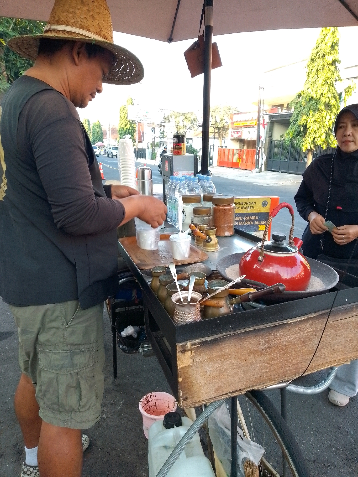 In Jember, Indonesia, a man wearing a straw hat and a dark t-shirt stands behind a mobile food cart. He is in the process of preparing a drink, with his hands holding a small white cup over a larger container. The cart itself is laden with various items, including bottles of water, jars of spices or powders, and a red teapot sitting on a bed of sand. To the right of the cart, a woman in a black hijab and traditional clothing is visible. She is holding what appear to be banknotes in her hands, suggesting a transaction is taking place. The background reveals a street scene with buildings, trees, and parked vehicles, indicating an urban environment. The overall scene depicts a street vendor operating their business on a typical day in Jember.