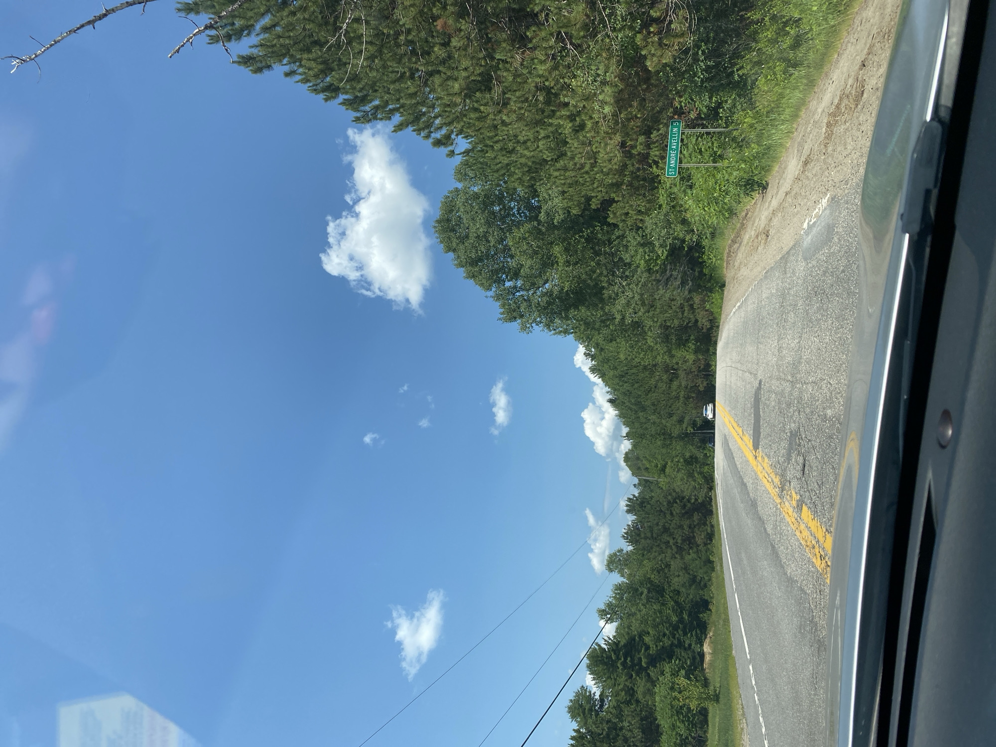 The image is a first-person view from a vehicle driving on a two-lane paved road under a bright blue sky with scattered white clouds. The road has a broken yellow center line and white lane markings. Dense green trees line both sides of the road, with taller coniferous trees on the right. A green street sign on the right side of the road reads "ST-ANDRÉ-AVELLIN 5". A white car is visible in the distance on the road. The foreground shows the dashboard and windshield of the vehicle. The environment appears to be rural, possibly in the region of Saint-André-Avellin, Canada, given the street sign.