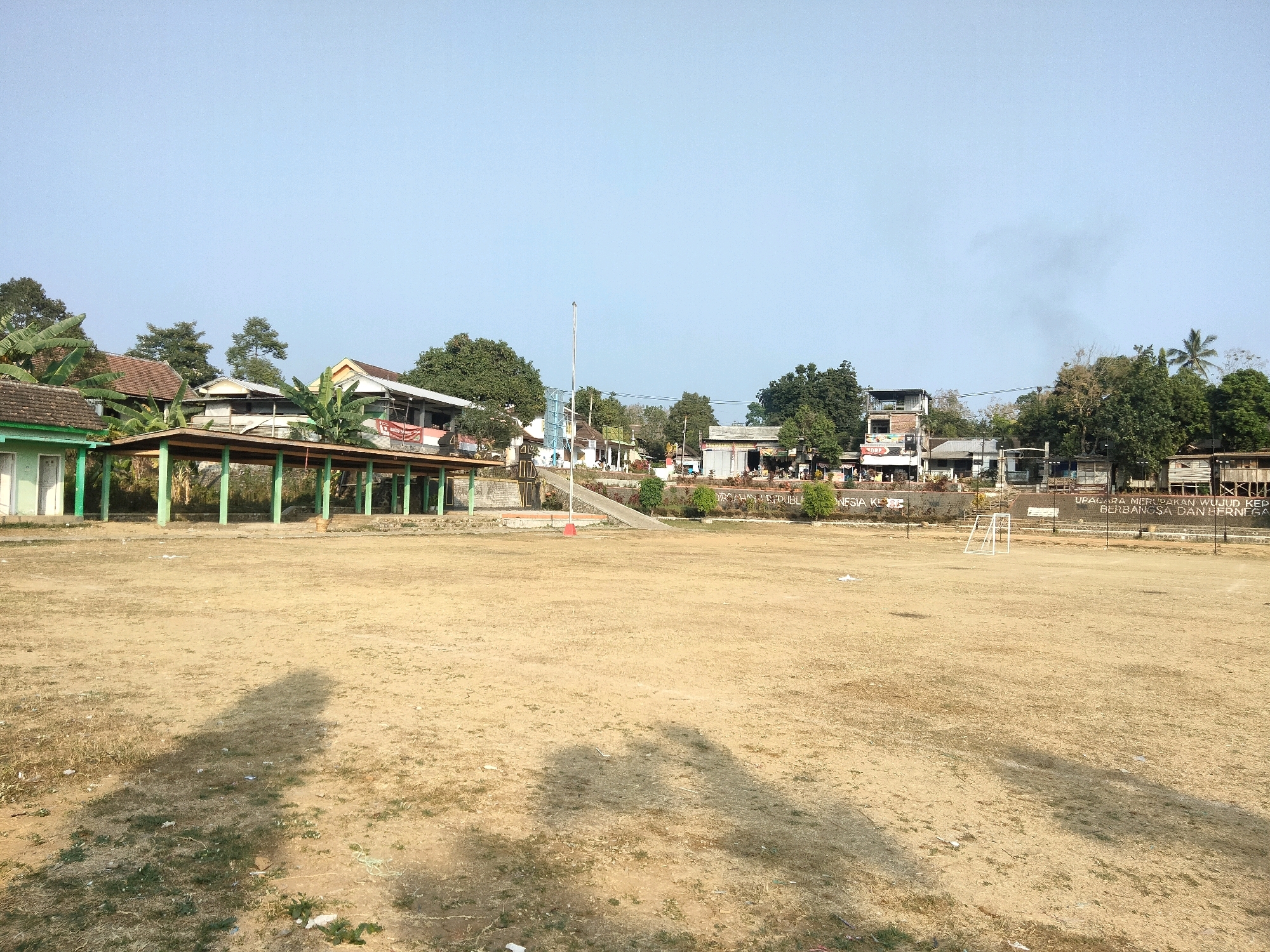 The image depicts an outdoor scene in Sawahan, Indonesia, likely a sports field or public gathering area. In the foreground, a wide expanse of dry, sparse grass covers the ground, with visible shadows cast by an unseen object or objects. The middle ground features a row of buildings and structures, characterized by a covered walkway with green posts and a corrugated metal roof. Behind this, several other buildings with varying rooflines and architectural styles are visible, interspersed with trees and vegetation. A flagpole stands prominently in front of some of these buildings. In the far background, more trees and a clear sky dominate the upper portion of the frame. There are no people visible in the image. The overall impression is one of a dry, rural or semi-rural environment.