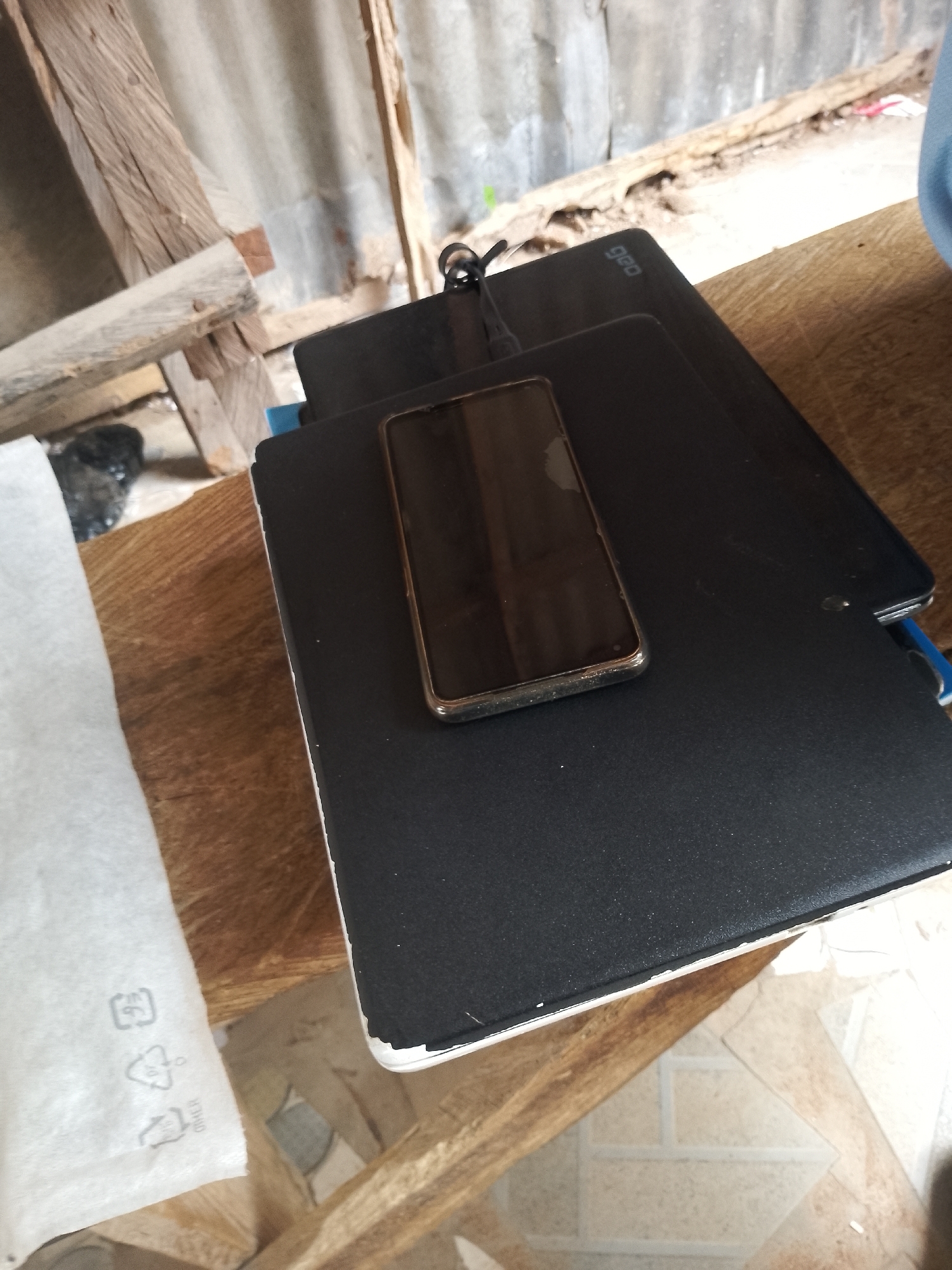 The image shows a close-up, high-angle view of a wooden surface. A black laptop is open, and a smartphone is placed on top of it. The smartphone's screen is dark. To the left of the laptop is a white, crinkled material with recycling symbols. In the background, there are some wooden planks and corrugated metal, suggesting an outdoor or unfinished indoor setting. The lighting suggests it is daytime. The location is identified as the city of Jalingo, Nigeria.