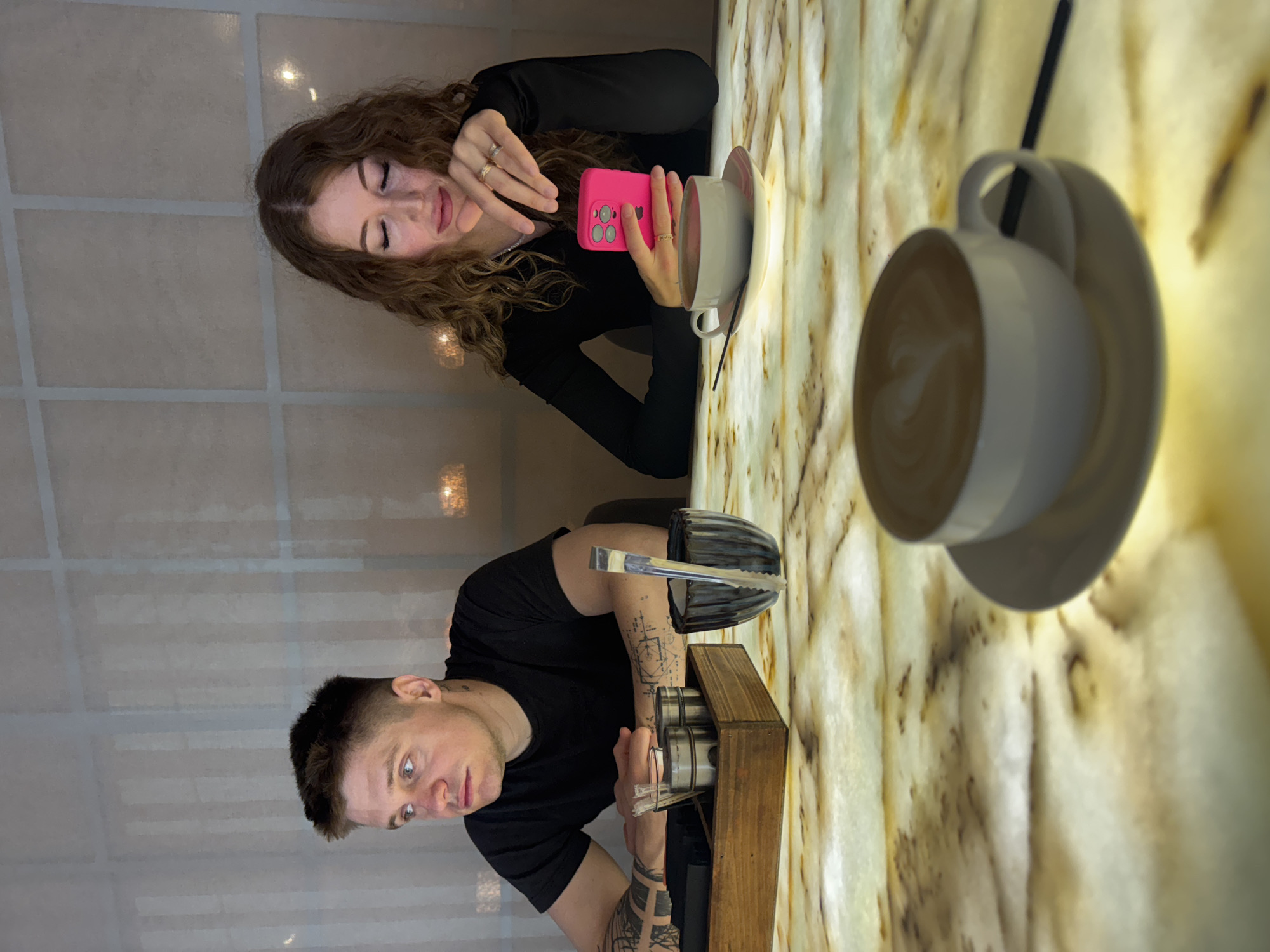 A woman with curly brown hair in a black long-sleeved shirt is holding a pink smartphone, seemingly taking a picture of a cup of coffee. Beside her, a man with short dark hair and tattoos on his arms is holding a silver object and appears to be interacting with a wooden box containing cylindrical metal containers. They are both seated at a table with a light beige and brown marbled surface. There is a second coffee cup and saucer nearby. The background includes a wall with a grid-like pattern. The location is implied to be in Sokhumi, Abkhazia, although no direct evidence in the image confirms this.