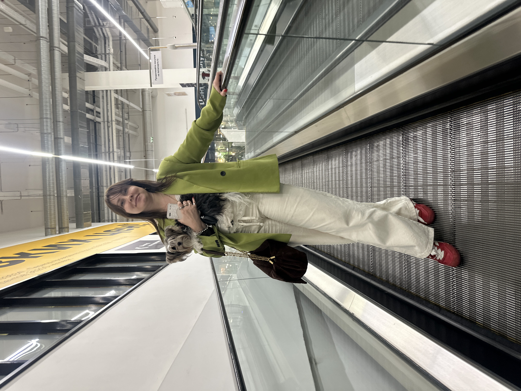 A woman stands on a moving walkway in what appears to be a retail or warehouse environment. She wears a green jacket, cream-colored pants, and red shoes. She holds a small dog in her left arm, which rests on a brown purse, and her right arm is holding up a cell phone in the air. The moving walkway is metallic, and there are industrial pipes and beams visible in the background. There is a sign in Russian, suggesting the location is in Russia, most likely Moscow.