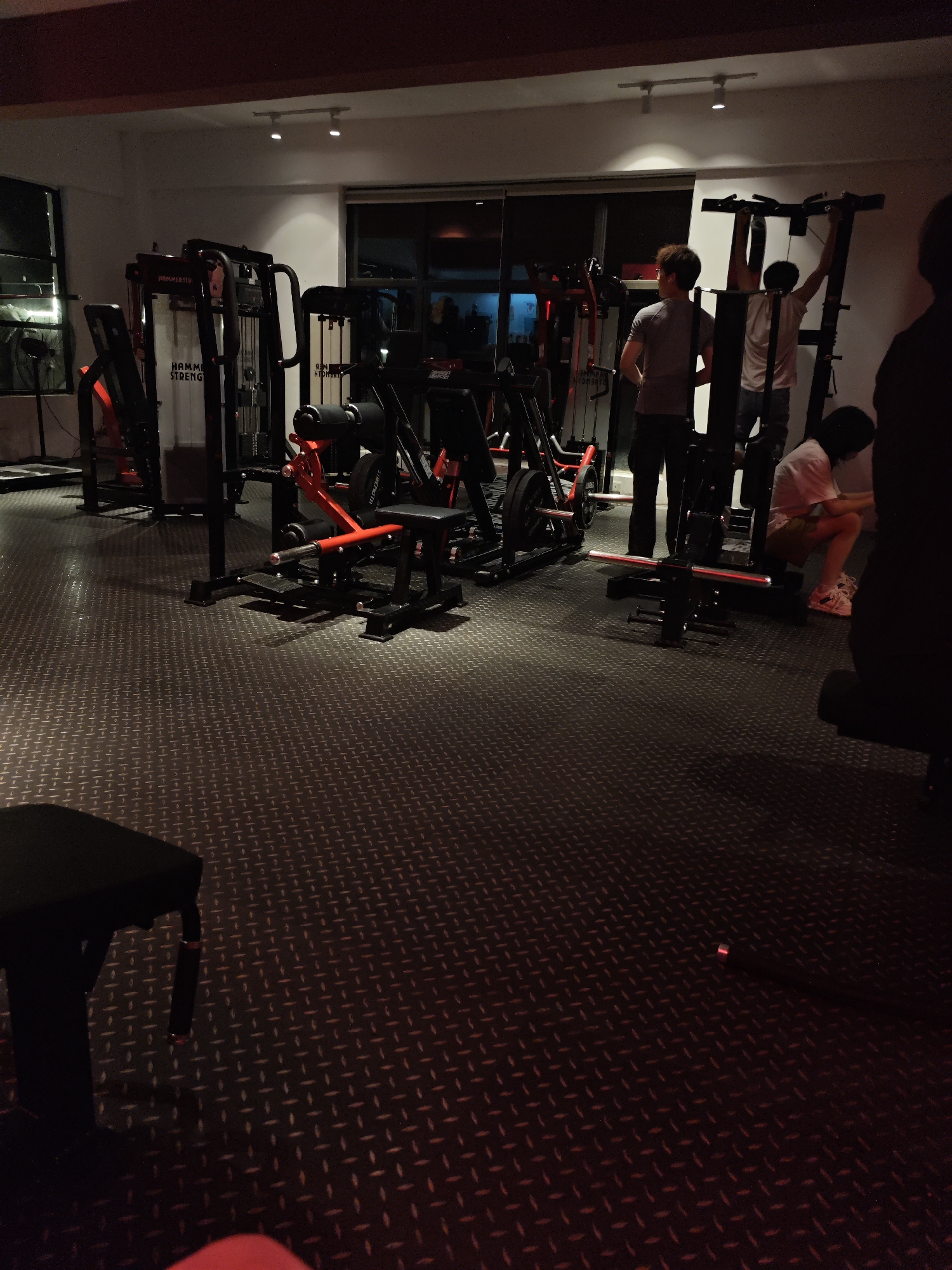 The image shows the interior of a gym. The floor is black with a textured, repeating pattern. Several pieces of gym equipment are visible, including weight machines with red and black accents. Three people are in the background: two males are standing and one is seated. One male is doing pull-ups on a pull-up machine. The gym has adequate lighting. The location is potentially in the Chongqing High-tech Zone, China.