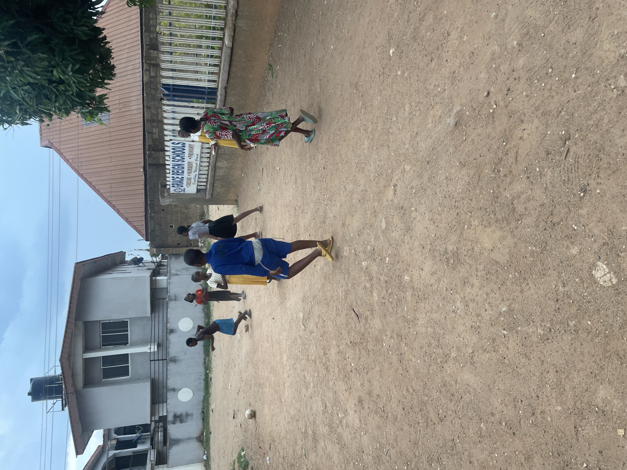 The image depicts an unpaved area adjacent to a building identified by signage as "Grace Reign Schools" with the words Creche, Nursery and Primary underneath the school name. Several individuals are present, appearing to be schoolchildren, some are actively engaged in playing a game, while others observe. One child is wearing a blue tracksuit and yellow footwear, bending over, while another child is wearing a blue outfit and on the ground. A ball is near them on the ground. A woman in a multicolored dress walks past the building. The surrounding environment includes trees, other buildings and utility poles, indicating an urban, likely residential area. Without additional information, determining its precise location in Jos, Nigeria is not possible.