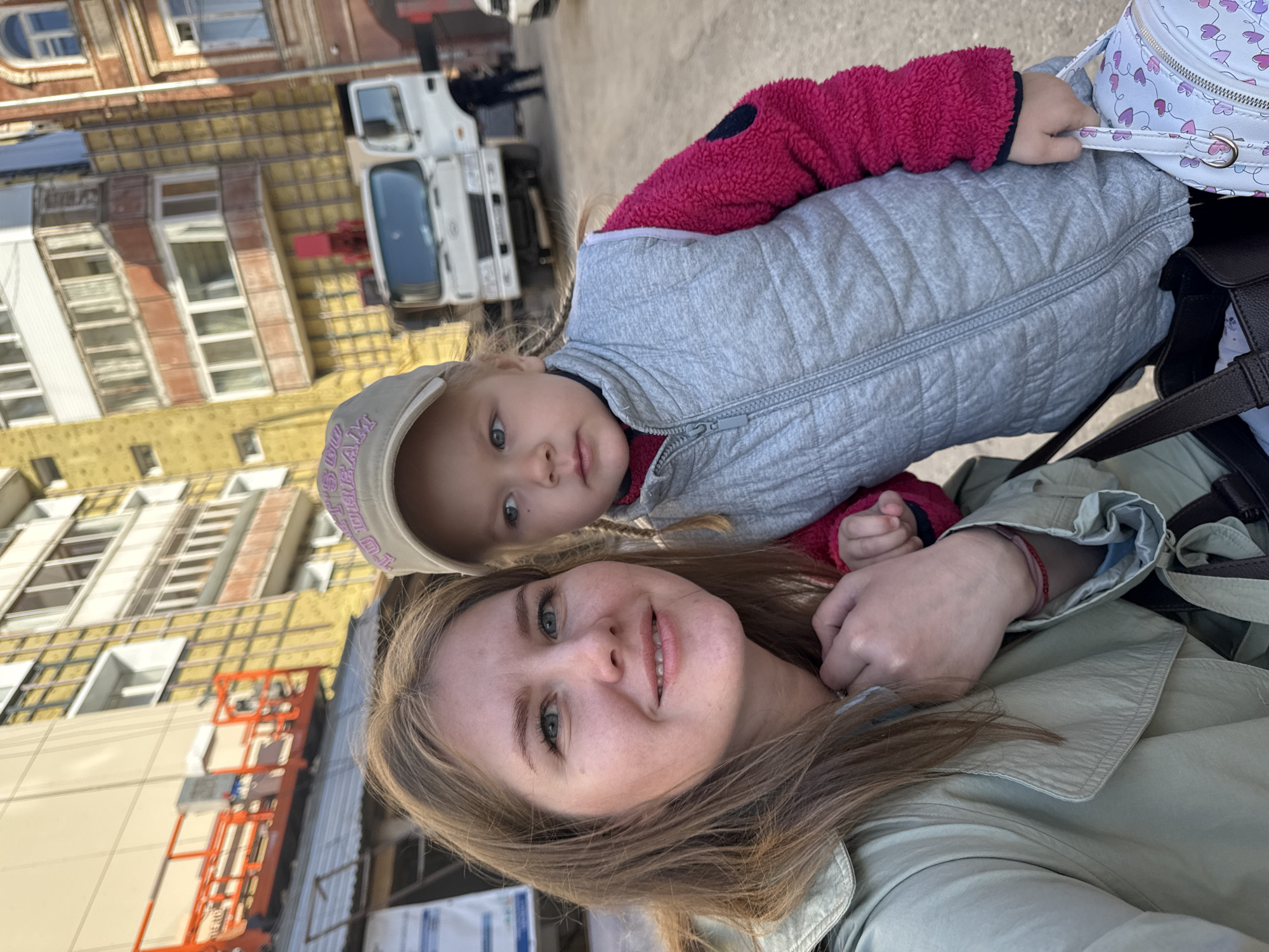 A heartwarming selfie captures an adult woman and a young child in an urban environment in Perm, Russia, bathed in bright daylight. The image, presented sideways, features the woman, likely the mother, with light brown hair and green eyes, offering a gentle smile to the camera. She wears a light green or khaki-colored jacket, with her arm appearing to support or embrace the child.
The child, a young girl with blonde hair and blue eyes, wears a light grey puffer vest over a fuchsia-pink long-sleeved top and a light-colored baseball cap with purple text that resembles "SEW FAM". Her expression is neutral and observant as she looks towards the camera. One of her hands is visible, resting on the woman's upper body, and she holds a small white backpack decorated with purple heart patterns.
The background reveals a bustling urban setting with buildings under construction or renovation, evident from exposed yellow insulation panels, scaffolding-like structures, and a parked white truck. The clear, bright conditions suggest a sunny daytime scene, highlighting the casual outdoor moment shared by the two individuals.