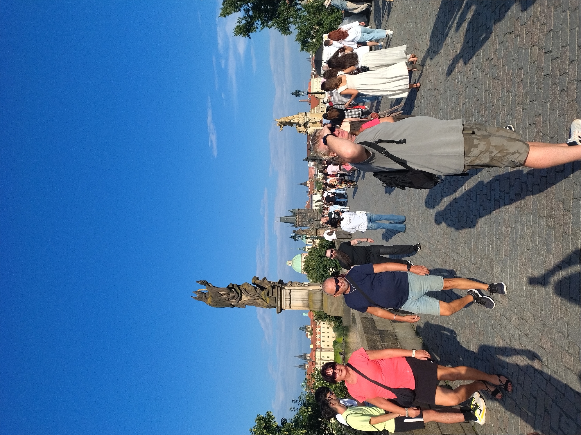 This is a vertical, eye-level, full-shot photograph taken on a cobblestone bridge in Prague, Czechia. The bridge is crowded with people walking in both directions. In the foreground, a man in a gray t-shirt and camouflage shorts is facing right, shielding his eyes from the sun. To his left, a couple in matching dark shirts and shorts are walking towards the camera. Behind them, a group of people, including women in a pink t-shirt and a black skirt, and a man in a dark blue t-shirt, are walking away from the camera.
In the background, there are several statues and architectural elements, including a large stone statue of a robed figure holding a torch. The city's skyline is visible, with buildings featuring domed roofs and spires. The sky is clear and bright blue. The overall impression is of a busy, sunny day on a historic bridge, likely the Charles Bridge, given the architectural style and the presence of statues.