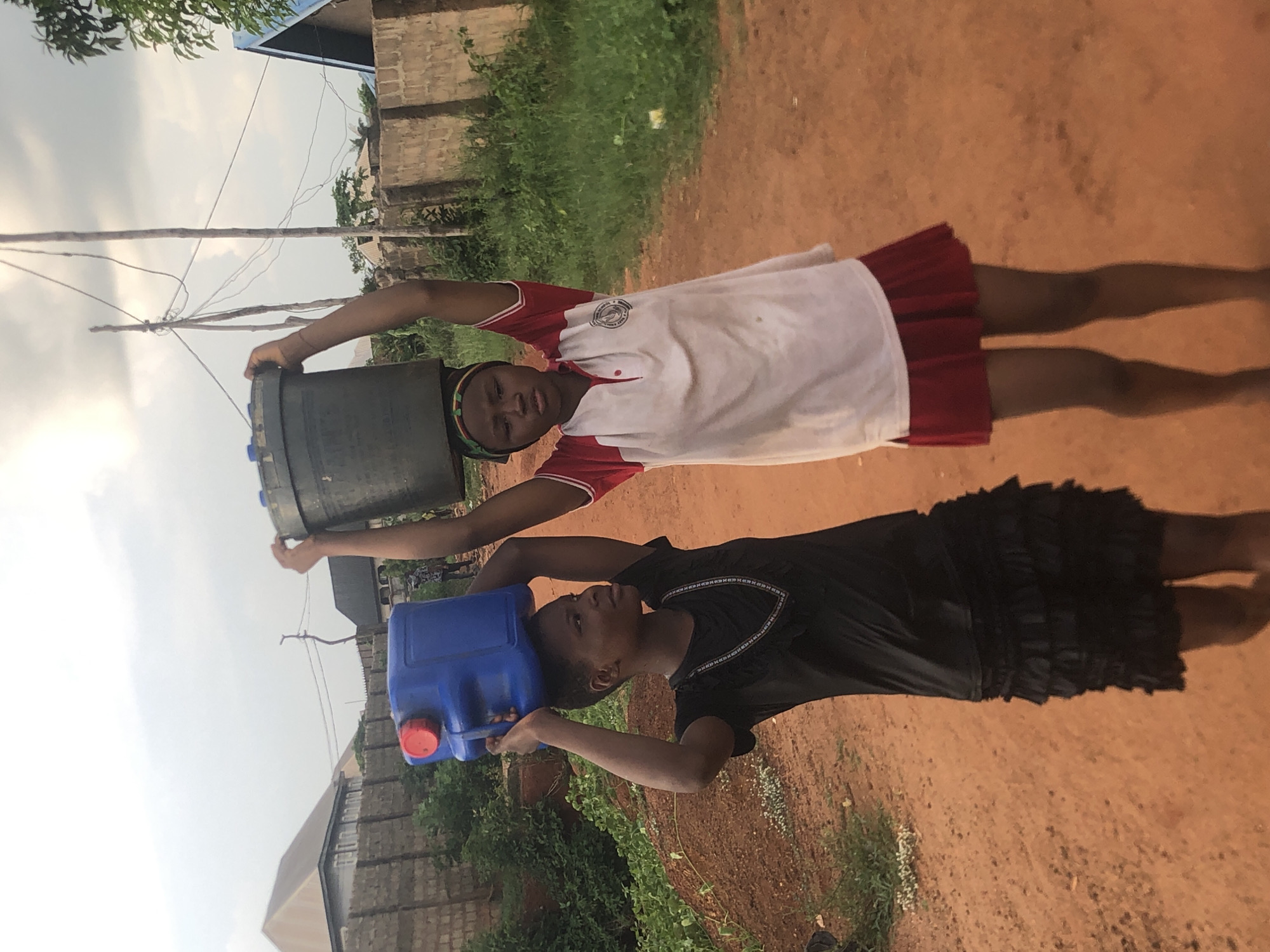 Two girls are carrying containers of water on their heads in Nkwelle, Nigeria. One girl wears a white shirt with red trim and a red skirt, while the other wears a black dress. They are walking on a dirt road with vegetation on either side. Buildings are visible in the background. The sky is overcast. The girl in white carries a large, gray container, and the girl in black carries a smaller, blue container.