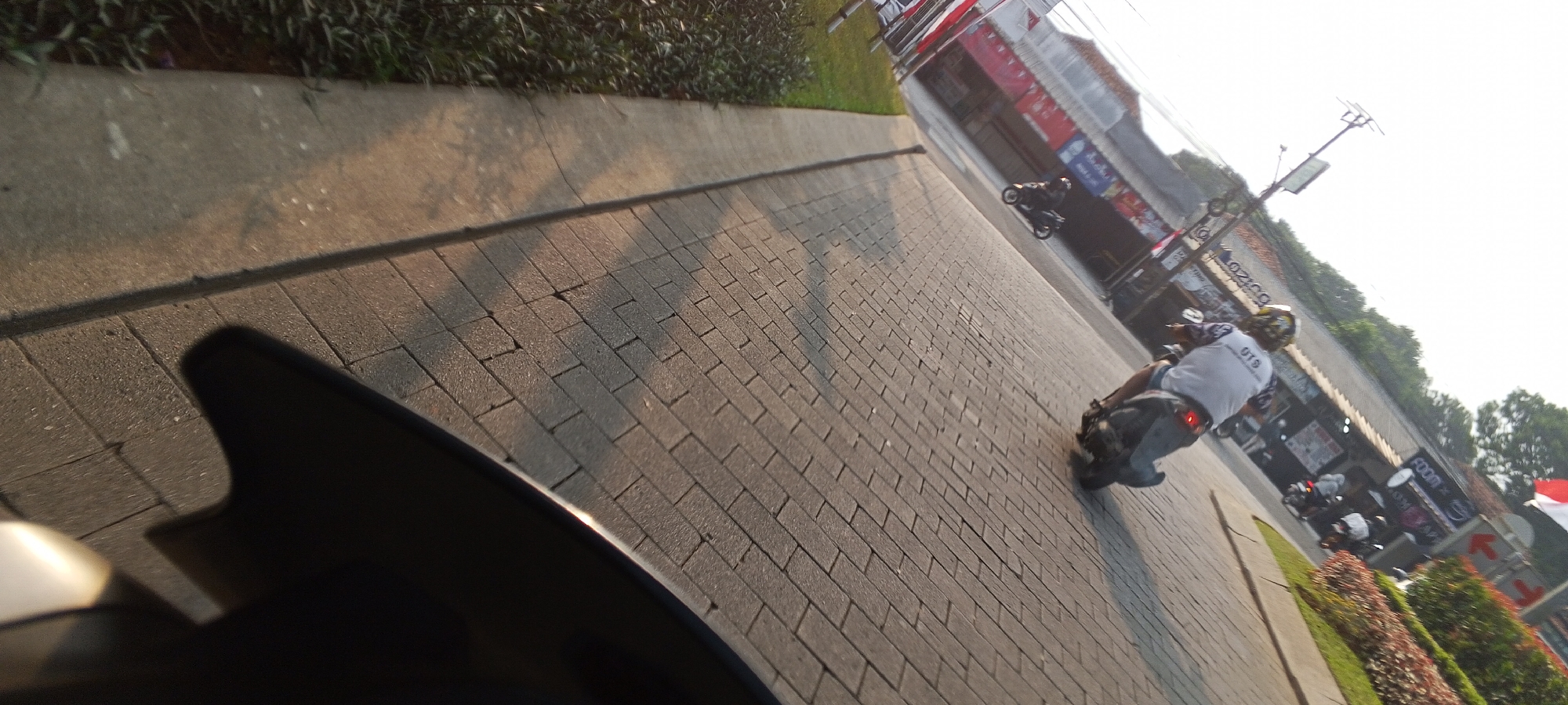 The image depicts a street scene in Depok, Indonesia, with a focus on a paved walkway and a road. The walkway is constructed of interlocking brick pavers, with a concrete curb separating it from a grassy area and dense green shrubbery. The road appears to be a paved surface where several individuals are riding motorcycles.
In the foreground, a portion of a dark-colored vehicle, possibly a motorcycle windshield, is visible on the left side of the image, casting a shadow on the walkway. Further down the walkway and road, two individuals are riding motorcycles. The motorcycle closer to the viewer has a rider wearing a white t-shirt with text on the back and a yellow helmet. This rider is captured in profile, with their motorcycle's brake light illuminated. Another motorcycle is visible further ahead, with its rider also wearing a helmet.
The background features buildings with storefronts and signage, suggesting a commercial area. Signs visible include "FOOM" and other illegible text. Red and white flags are also present on some of the buildings. Trees and foliage are seen behind the buildings, indicating a presence of greenery within the urban environment. The overall lighting suggests it is daytime, with the sun casting long shadows. The image is taken from a low angle, possibly from another moving vehicle.