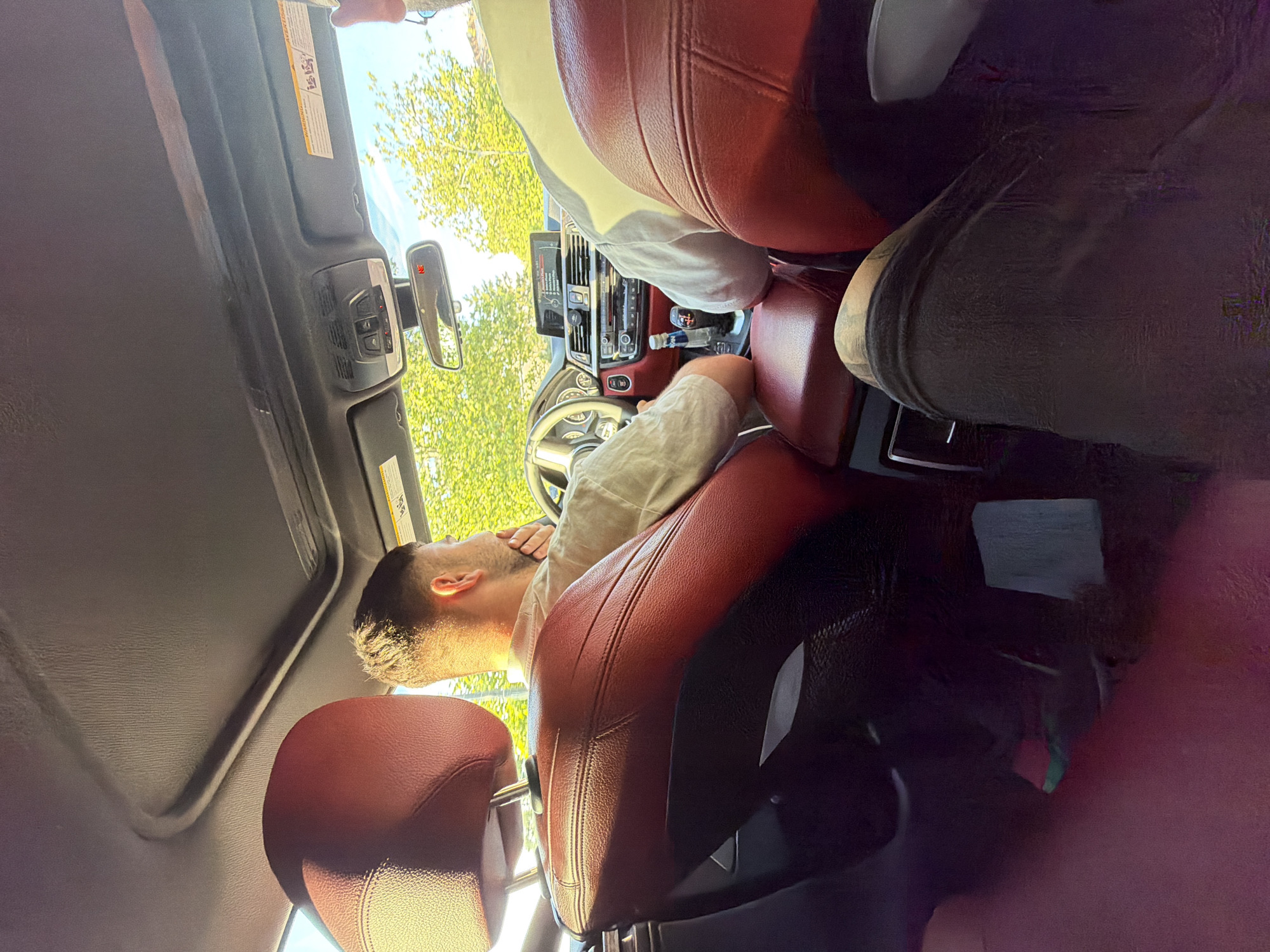The image shows the interior of a car with two people. The person in the driver's seat is a male with short hair, wearing a light-colored shirt. He is looking to his right, with his right hand on his chin. The car has red leather seats and a sunroof. Outside the car, trees and a building are visible. The image was taken in Ruse, Bulgaria.