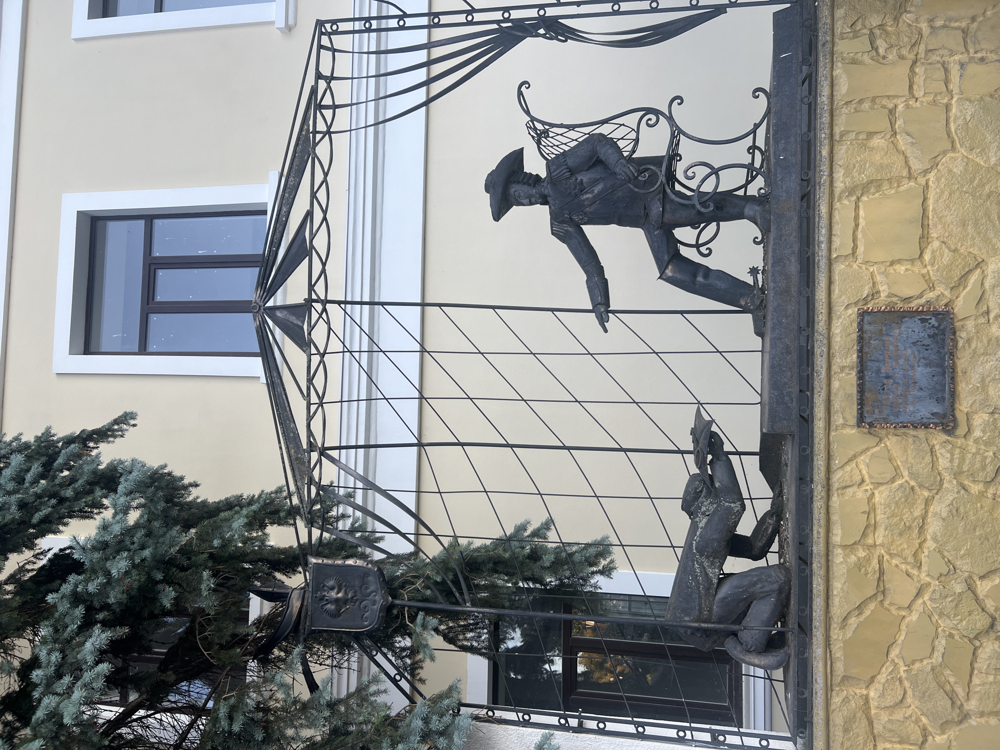 This is a metal sculpture located in Bender, Moldova, depicting two figures within a decorative metal enclosure. On the right, a man in historical attire, including a tricorn hat, is seated on an ornate chair, pointing to the left. On the left, a man is kneeling, appearing to be either writing or presenting something. The sculpture is mounted on a stone wall, and a portion of a large evergreen tree is visible on the left side of the frame. A coat of arms is also visible on a metal post to the left of the enclosure. The sculpture is set against the backdrop of a building with windows.