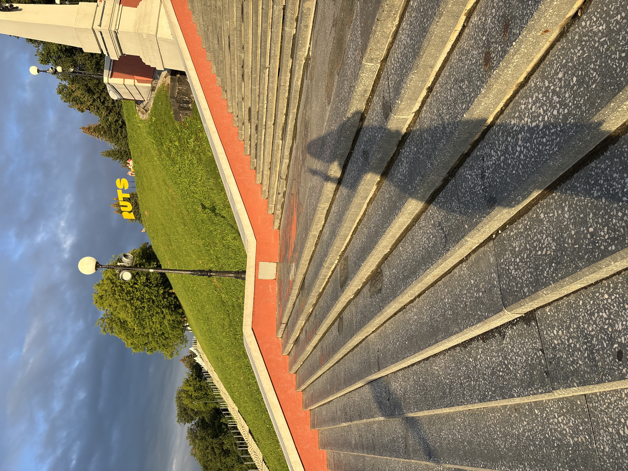 The image shows a wide stone staircase leading up a grassy slope in Nizhny Novgorod, Russia. The staircase is made of grey, speckled concrete with red trim along the sides. The sun is setting, casting long shadows across the steps and illuminating the grass on the slope. At the top of the slope, large yellow letters spell out "RUTS". A shadow of a person holding a phone is visible on the stairs in the foreground. In the background, a dark storm cloud is visible in the sky.