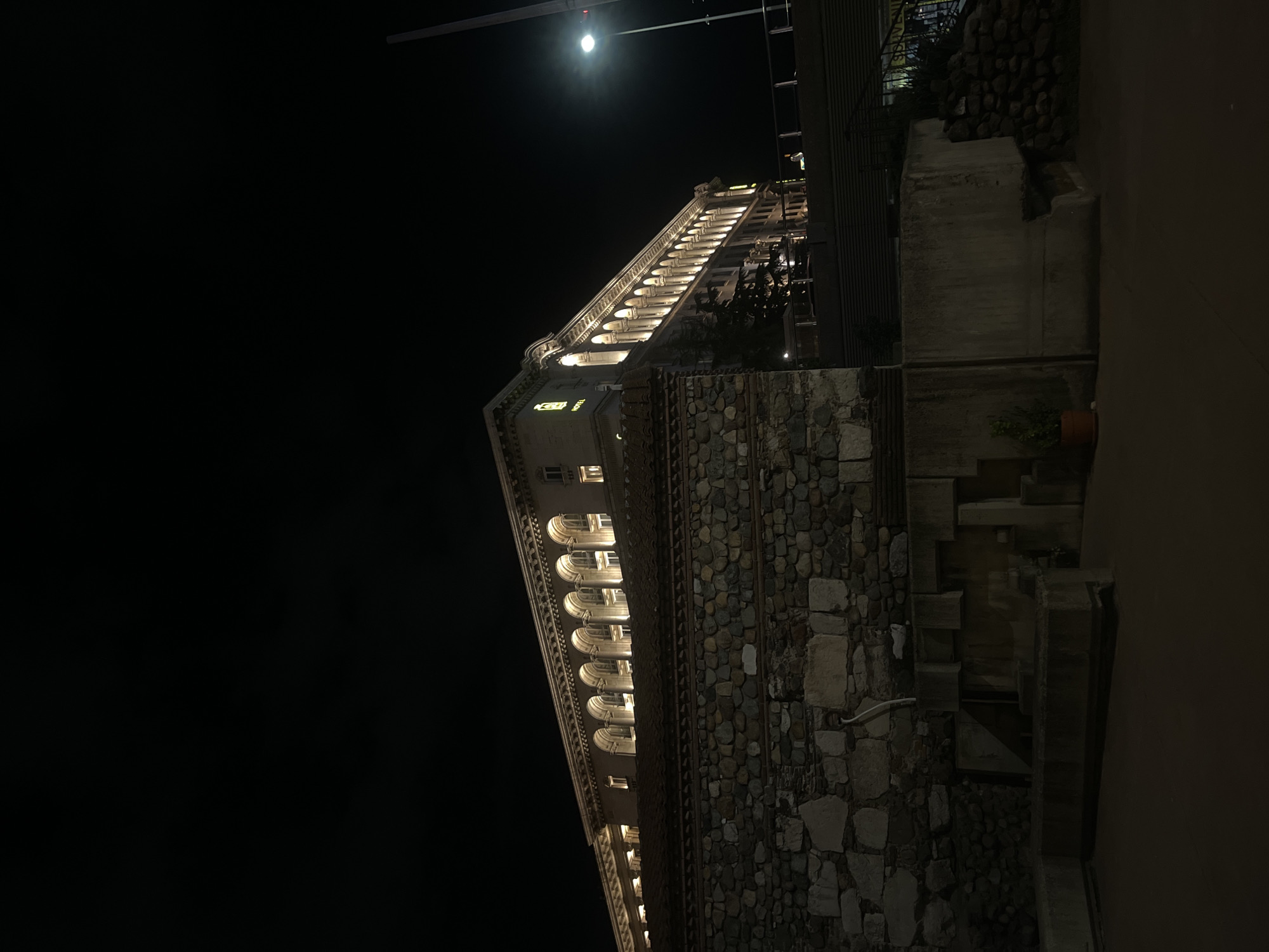 A low-angle, night-time shot features a tall, lit building identified as a hotel via illuminated signage, positioned against a dark sky with a visible moon. The building has a stone facade up to a certain height, transitioning to a white, multi-storied structure with arched windows and illuminated terraces. The architectural style includes classical elements such as balustrades and decorative cornices. To the right of the building, a terraced stone wall and landscaping elements are visible. The scene appears to be located in an urban setting, likely Sofia, Bulgaria.