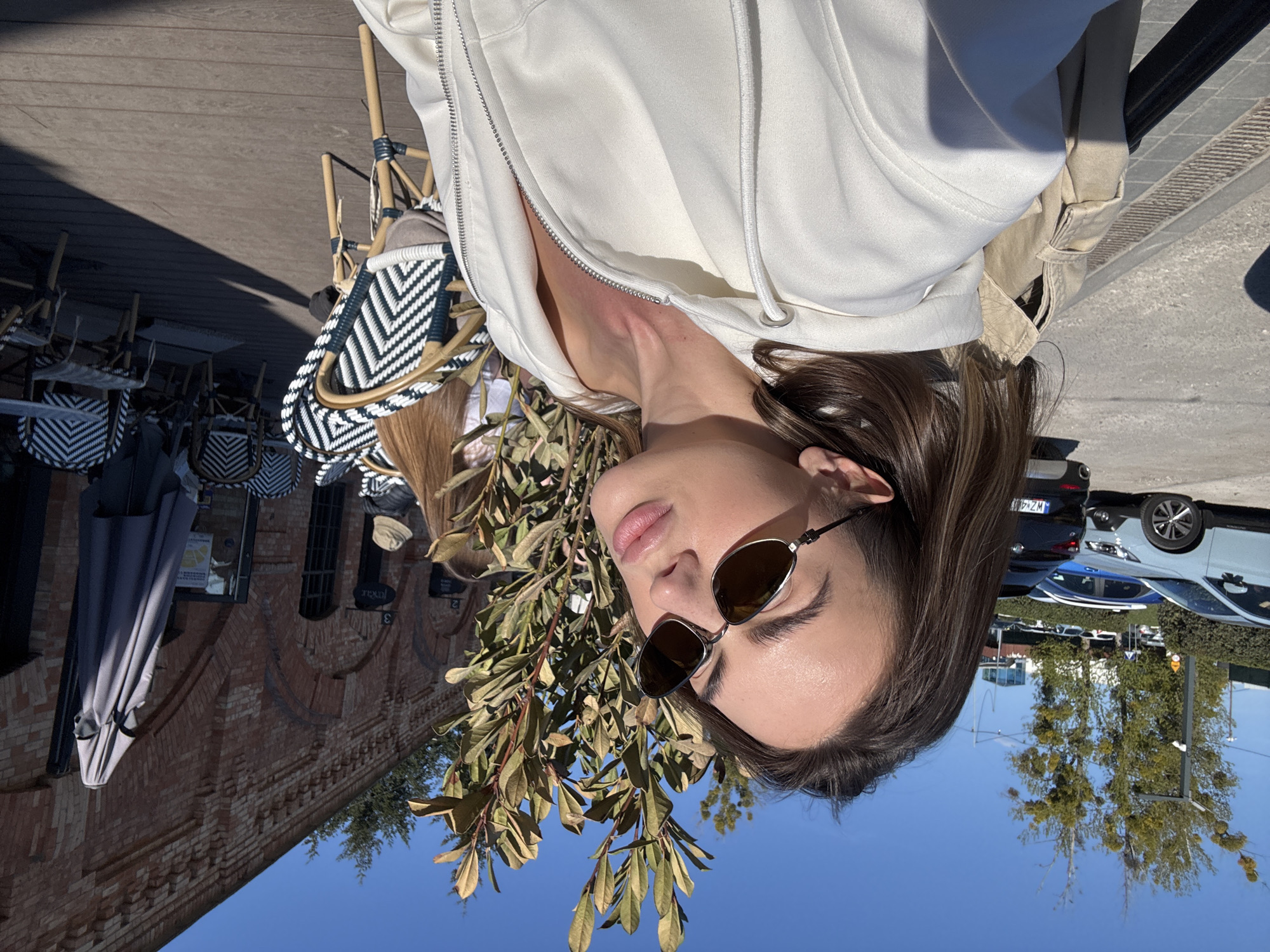The image presents an outdoor scene, likely a cafe or restaurant patio, on a bright, sunny day in what appears to be Warsaw, Poland. The primary subject is a person, possibly female, seen from the chest up, positioned slightly to the left of the frame. They are wearing a white hooded zippered sweatshirt and dark-lensed sunglasses, with their brown hair visible. Their expression is neutral and relaxed, and they are looking towards the right side of the frame. A leafy branch with brown-green leaves extends prominently across the foreground, partially obscuring the person.
In the background to the left, several black and white patterned wicker chairs and small tables are arranged, indicative of an outdoor seating area. A bag with a similar black and white pattern rests on a chair behind the person. Further back, a multi-story brick building with arched window details is visible, adding an architectural element to the setting. A grey, folded outdoor umbrella is situated near the building. On the brick facade, a sign displaying "PAXAR" and the numbers "2" and "3" can be seen.
To the right and in the far background, an urban environment unfolds with parked cars, one of which has a partially visible Polish license plate starting with "WZ". Beyond the cars, several tall trees and a light pole are discernible under a clear, blue sky. The scene is well-lit by natural sunlight, suggesting a pleasant daytime setting. The overall impression is one of leisure and relaxation in an outdoor, city environment.
