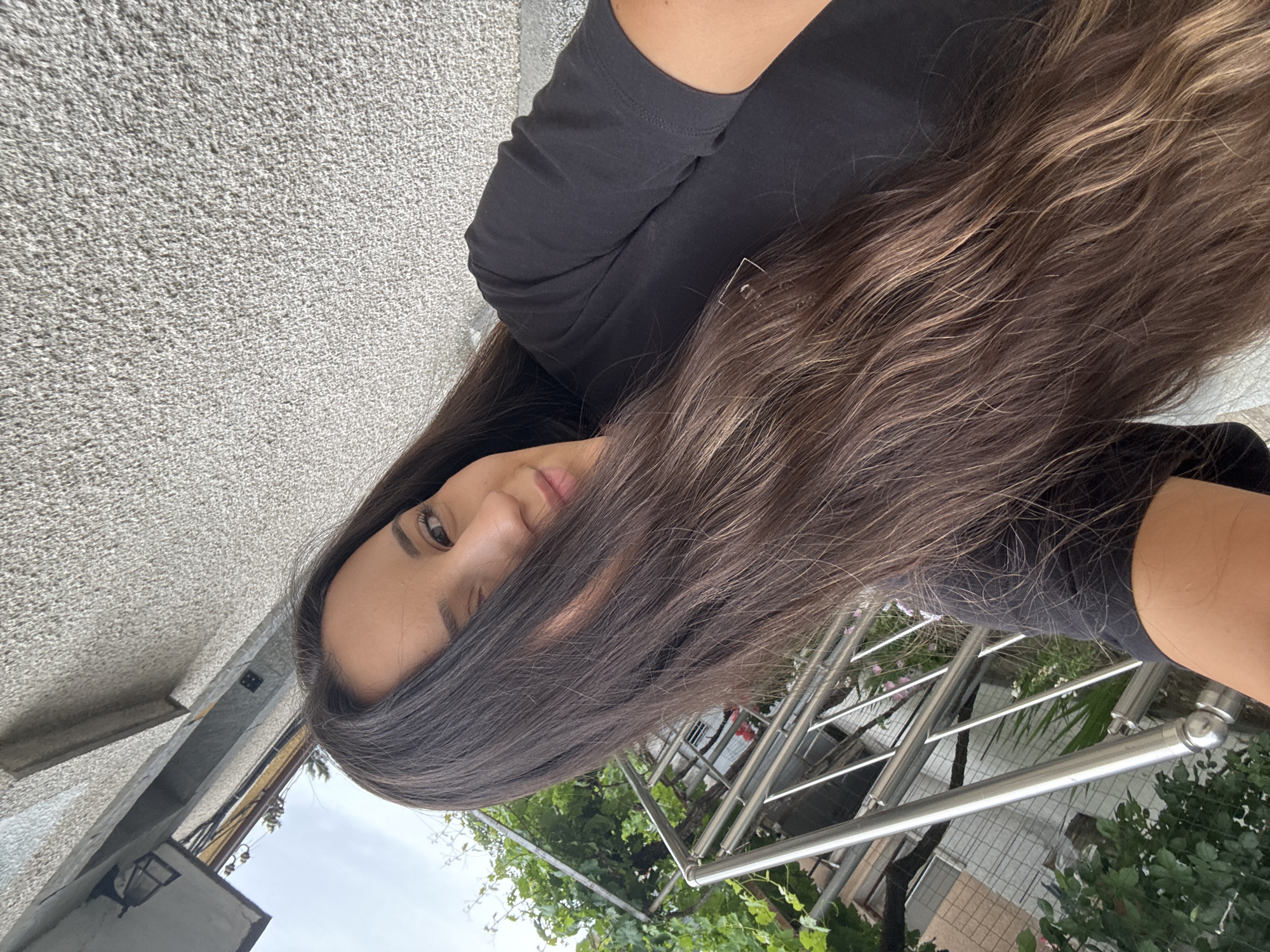 An overhead, outdoor, eye-level shot captures a woman in a black t-shirt leaning forward and looking towards the camera. Her long, brown hair covers the left side of her face. She is outside, with a grey textured wall behind her and a metal railing and lush green plants to her left. The lighting is overcast, suggesting a cloudy day.