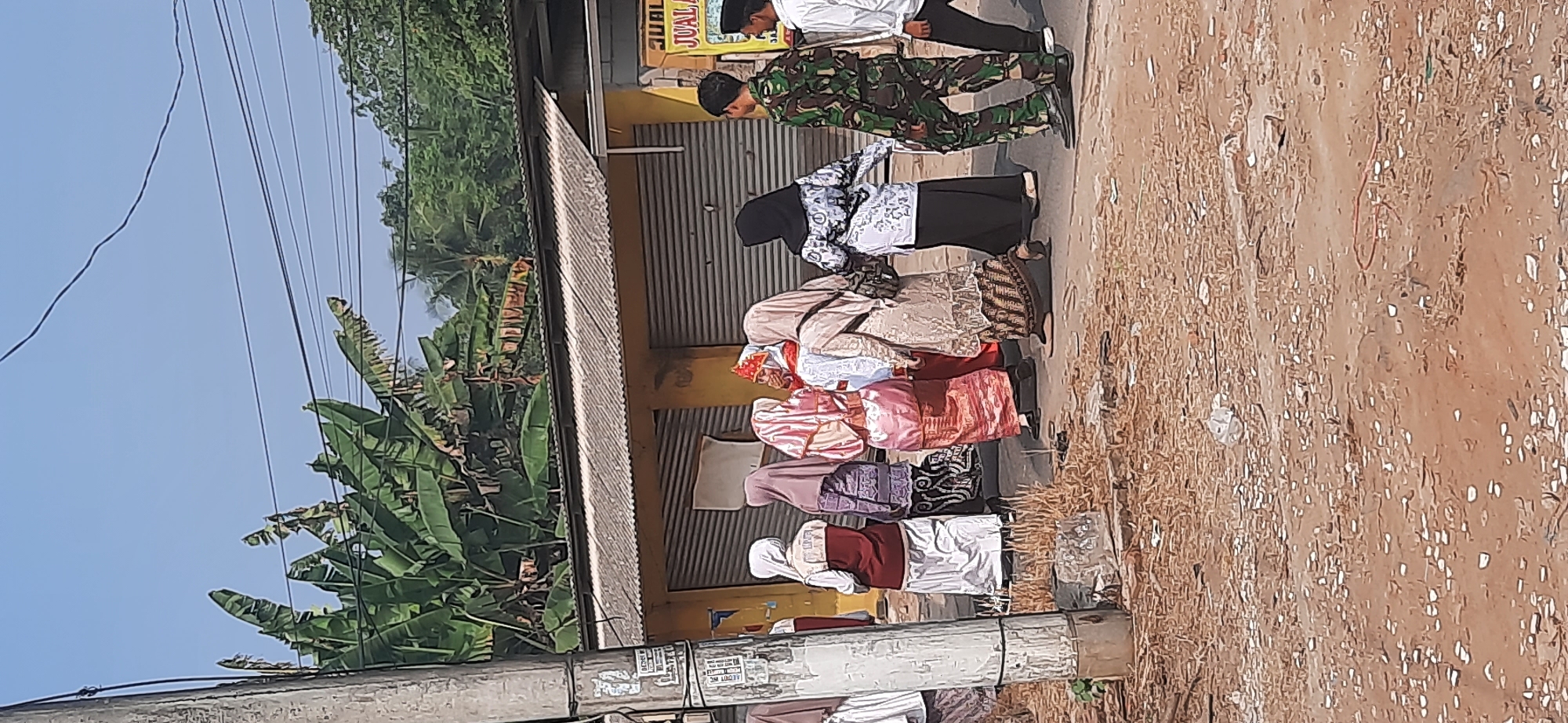In Cisauk, Indonesia, a group of seven individuals walks along a dirt path beside a yellow building with a corrugated metal roof. The group consists of five women and two men. Four of the women are wearing headscarves and long dresses, one with a traditional red and gold patterned outfit, and another in a white and patterned outfit. One woman is wearing a black headscarf and black patterned top with a black skirt. The two men are dressed in military camouflage uniforms. The building appears to be a commercial establishment with closed roll-up doors. Banana trees are visible in the background, and a utility pole stands to the left of the frame. The ground is covered in dirt and scattered debris. The sky is clear and blue.