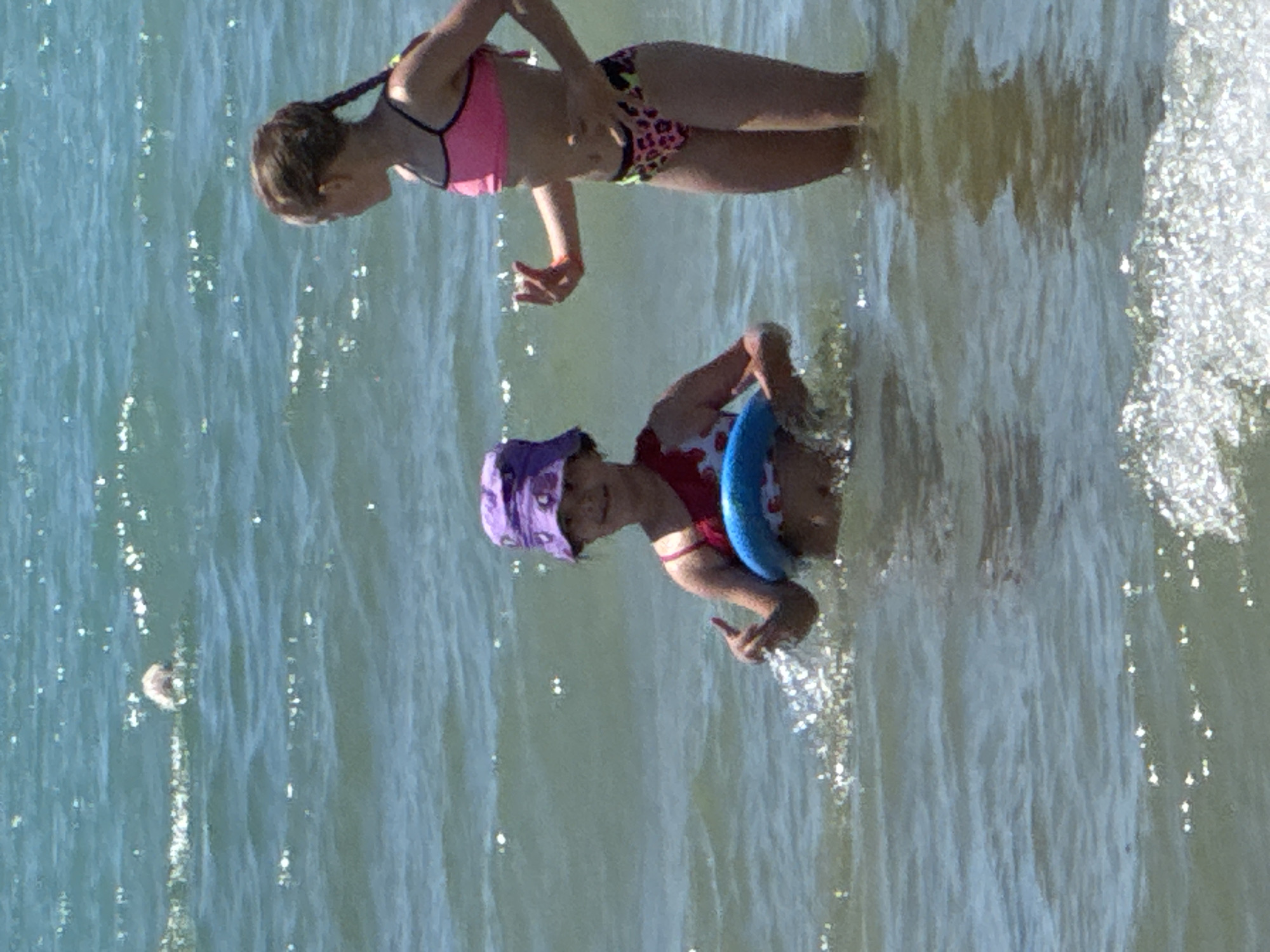 The image shows two young girls in a body of water. The girl in the foreground is wearing a red swimsuit and a purple swim cap. She is holding a blue inflatable ring. She is smiling and looking towards the viewer. The girl in the background is wearing a pink bikini and leopard print bottoms. She is also in the water. The water appears to be shallow and clear, with sunlight reflecting off the surface. The scene is likely a beach or a lake on a sunny day. The overall mood is joyful and playful, suggesting a fun summer day.