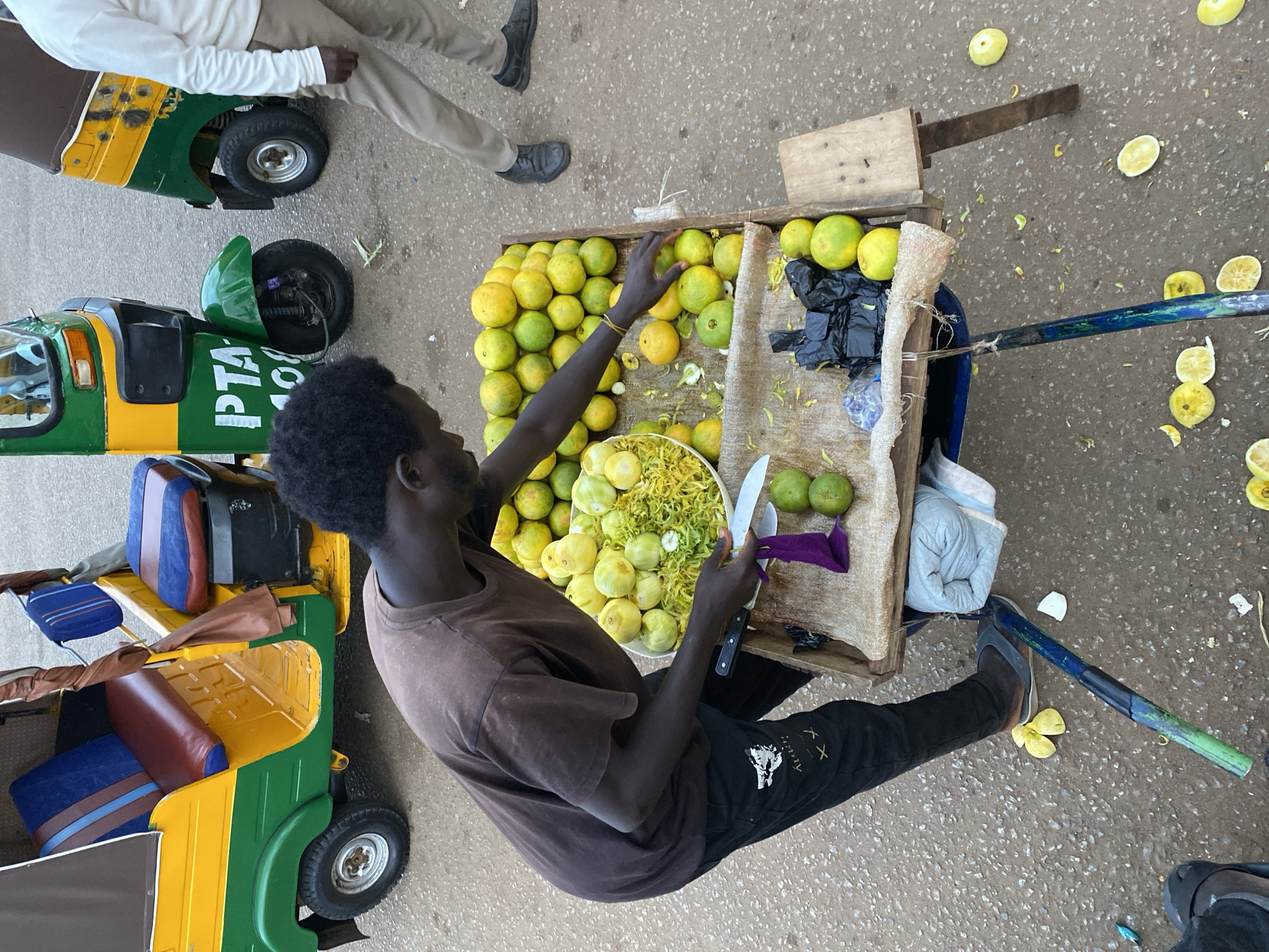 A young Black man with short, dark, curly hair, wearing a brown t-shirt and black pants with white graphic designs, stands behind a fruit stand in Bukuru, Nigeria. He is holding a knife in his right hand and a second knife is resting on the stand next to a purple cloth and some limes. The stand, made of wood, is on wheels and is laden with a large pile of green and yellow citrus fruits, some of which appear to be oranges. There is also a bowl of finely shredded green material, possibly fruit peelings or zest, and several whole limes. To the right of the fruit stand, a person in a white long-sleeved shirt and tan pants walks away from the camera. In the background, two yellow and green auto-rickshaws, commonly known as 'keke napep', are parked. One has "PTA 108" written on its side. The ground is paved with asphalt. The scene appears to be an outdoor market or roadside stall.