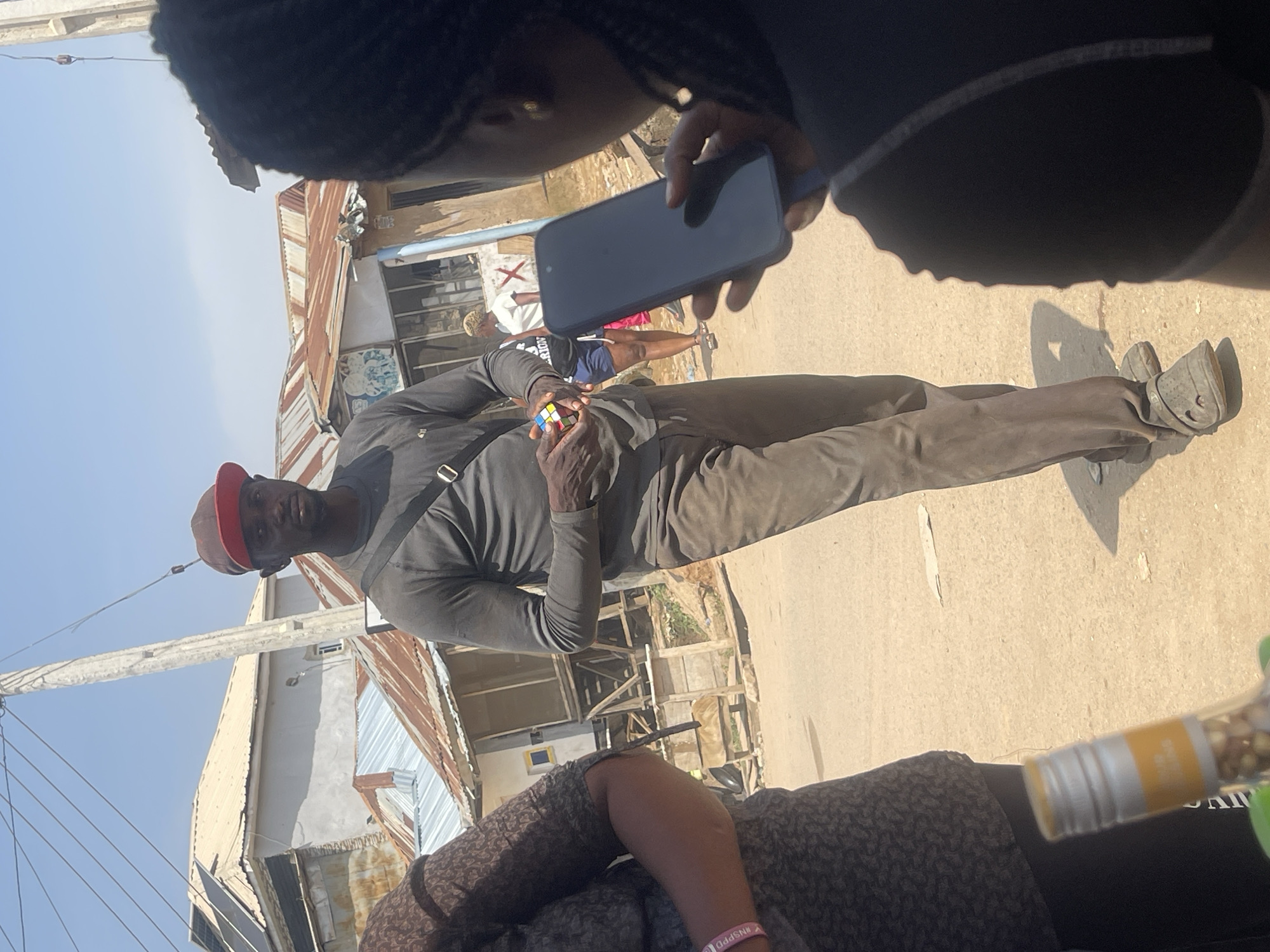 The image shows a man in a grey long-sleeved shirt, brown pants, and a red and brown baseball cap holding a Rubik's Cube. He is standing on a dirt road in front of buildings with corrugated metal roofs, typical of informal settlements in areas like Abuja, Nigeria. To his right, a person is holding a smartphone, seemingly recording or photographing the man. Another person, partially visible on the left, is wearing a patterned dress and a pink wristband. In the foreground, the neck of a bottle with a yellow and silver label is visible. The environment suggests a sunny day in a developing urban area.