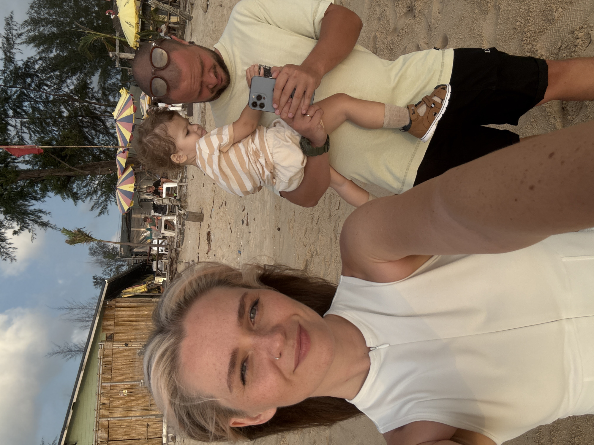 A medium shot captures a family of three on a sandy beach in Choeng Thale, Thailand. The foreground is dominated by the face of a woman with blonde hair, freckles, and a nose ring, wearing a white sleeveless top. To her right, a man with a beard and sunglasses perched on his head holds a young child. The child, wearing a striped t-shirt and light-colored shorts, looks towards a smartphone the man is holding. In the background, a building with a bamboo facade and thatched roof is visible, along with beach umbrellas and other patrons sitting at tables. The environment suggests a relaxed, casual atmosphere.