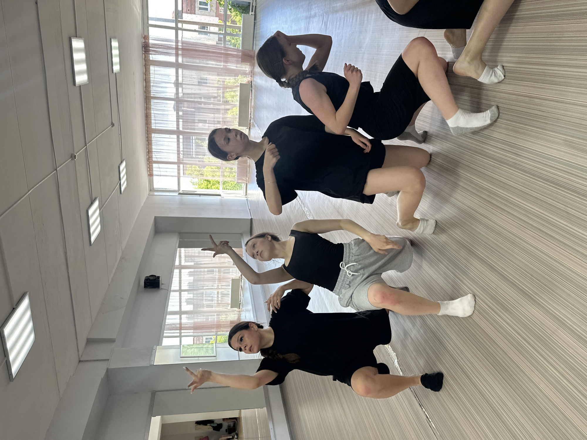 In a brightly lit dance studio with a wood-like floor and large windows with sheer curtains, five young women are practicing a choreographed movement. Four are in the foreground, and one is partially visible on the far right. The women are wearing casual black or grey athletic clothing and socks. Two women are kneeling on their right knees with their left legs extended. Another is kneeling on both knees, and the fourth is in a deep lunge. The two women on the left have their arms raised, with fingers pointed upwards, in a gesture of reaching or grasping. The environment includes overhead fluorescent lighting panels and mirrors on the left wall. The scene appears to be a contemporary dance class. The location of this dance studio is Votkinsk, Russia.