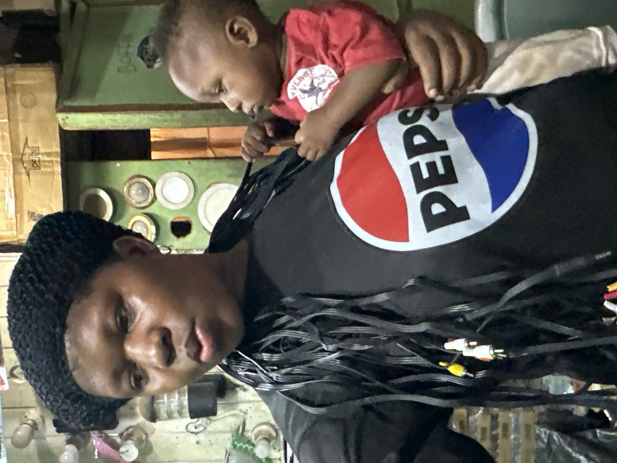 In Narayi, Nigeria, a woman wearing a black beret and a black t-shirt with a Pepsi logo stands facing forward. Her hair is styled in thin, black braids that hang down her chest. She has a child in her arms, who is wearing a red t-shirt. The background appears to be an indoor setting with shelves holding various items, including plates on the wall and boxes stacked. The lighting suggests it is daytime.