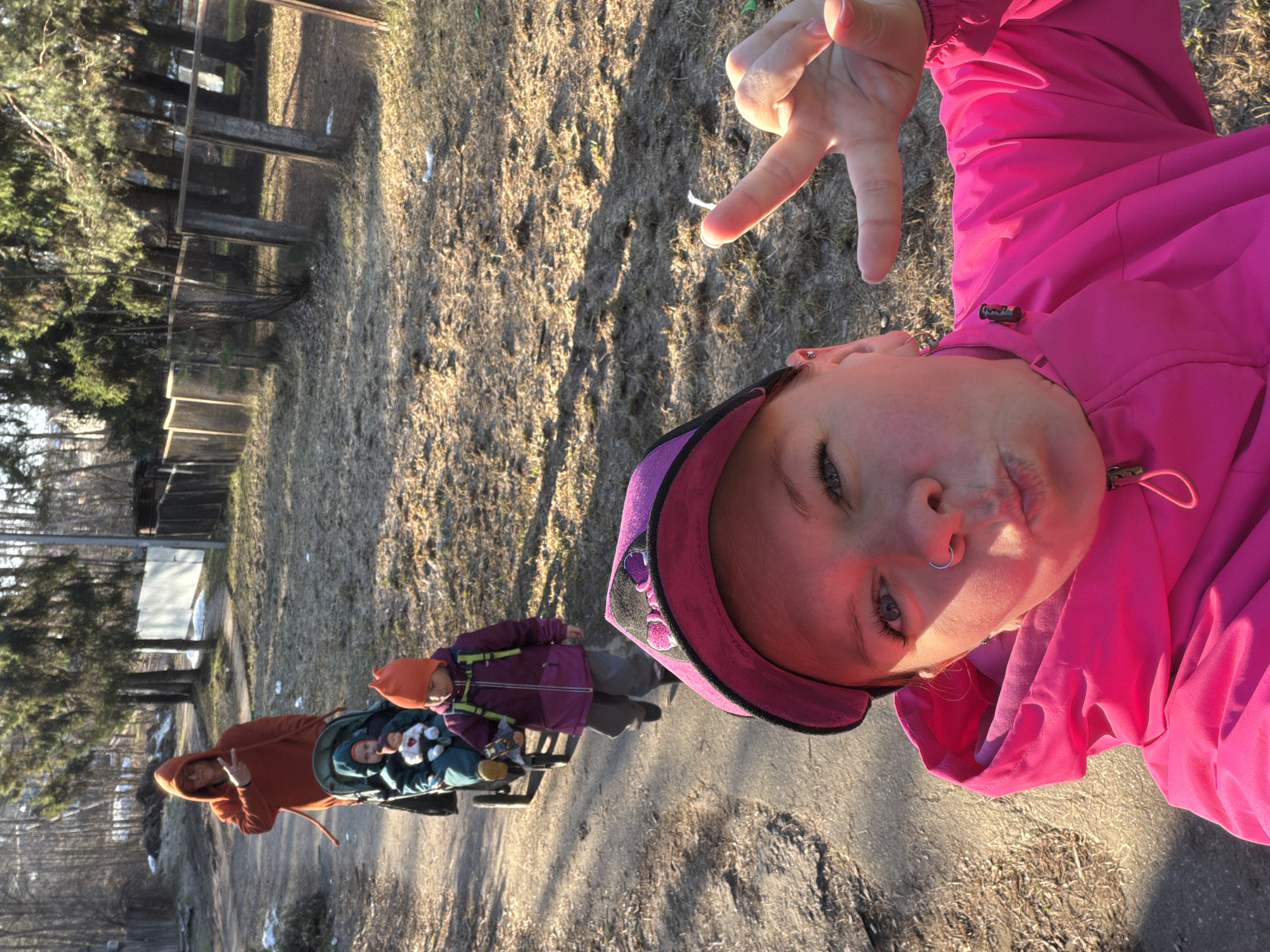 A selfie taken outdoors captures a young person in a pink jacket and purple cap making a peace sign, looking directly at the camera with blue eyes and a nose ring. The light catches their face, creating a warm glow. In the mid-ground, slightly out of focus, another individual wearing an orange hooded jacket also displays a peace sign while pushing a stroller containing two small children. A third child, dressed in a purple jacket and orange hat, stands next to the stroller.
The scene is set on what appears to be a dirt or gravel path, flanked by bare ground and patches of dry grass. Tall trees with sparse foliage line the background, suggesting late autumn, winter, or early spring. Several wooden or concrete structures, possibly benches or fences, are visible further back among the trees. The lighting indicates either early morning or late afternoon (golden hour), with long shadows cast across the ground by the strong sunlight. The overall atmosphere seems casual and playful. There is no visible text in the image. The setting is an outdoor area in Chaikovsky, Russia, possibly a park or wooded trail.
