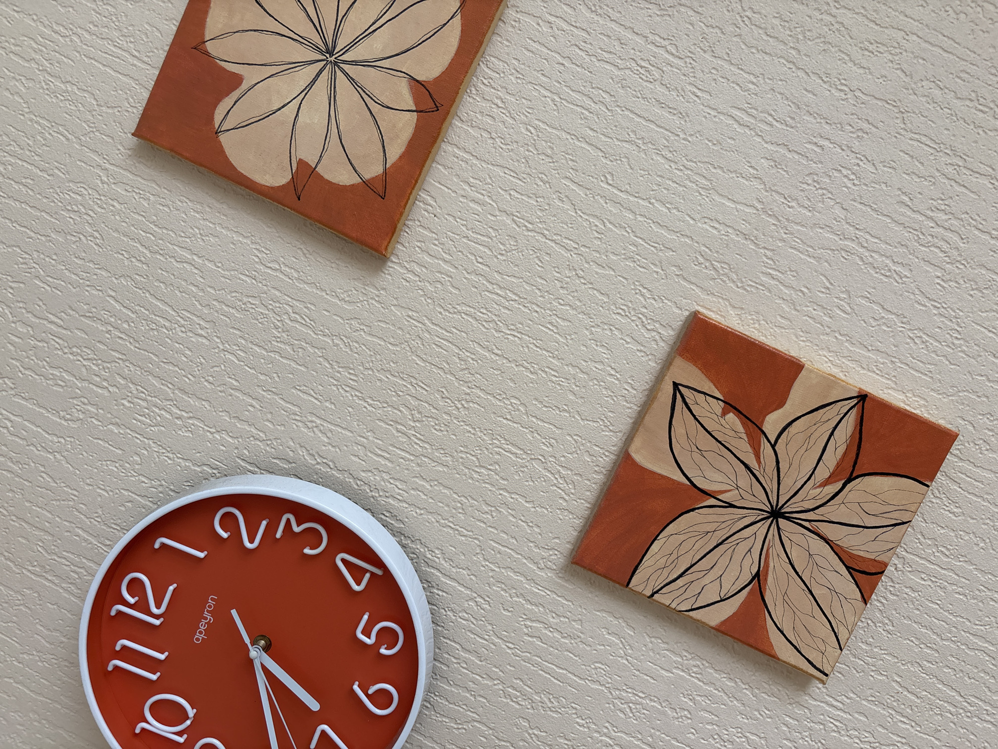 A bright orange wall clock with white numbers and hands is mounted on a textured, cream-colored wall. Two square canvases with abstract floral designs are also displayed on the wall. One canvas is positioned below and to the left of the clock, while the other is above and to the right. The floral designs feature a star-like pattern with dark outlines on a background of orange and beige. The image does not contain any information about the location in Krasnodar, Russia.