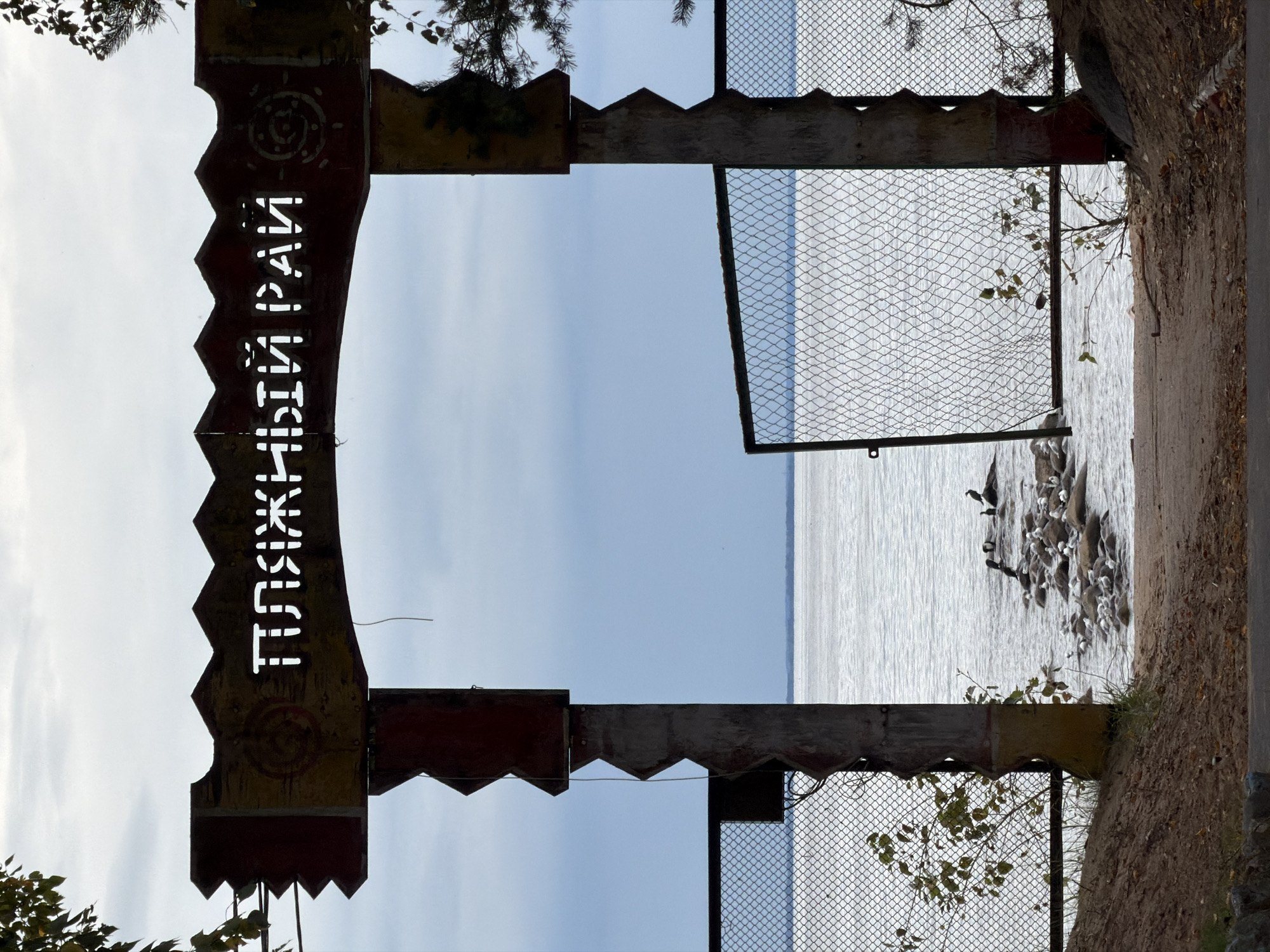 An outdoor, medium shot captures an entranceway to a beach in Komarovo, Russia, under a cloudy sky. The entrance is framed by two weathered, wooden posts and a jagged, wooden archway. The archway features Cyrillic lettering that reads "ПЛЯЖНЫЙ РАЙ," meaning "Beach Paradise." The archway is adorned with painted symbols, a circular pattern on the left and a sun-like symbol on the right.
To the left and right of the entrance, chain-link fences extend towards the water, creating an enclosure. The fence on the left is partially obscured by a small, leafy bush. The fence on the right has one gate open, revealing the path forward.
Beyond the fences and the entrance, a body of water, likely the Gulf of Finland, stretches to the horizon, its surface rippled by a gentle breeze. The water is dotted with many rocks, on which several seagulls and dark-colored birds are perched. The ground leading into the entrance is a sandy path, scattered with fallen leaves, indicating an autumn season. The overall impression is of a somewhat rustic and possibly abandoned beach entrance.