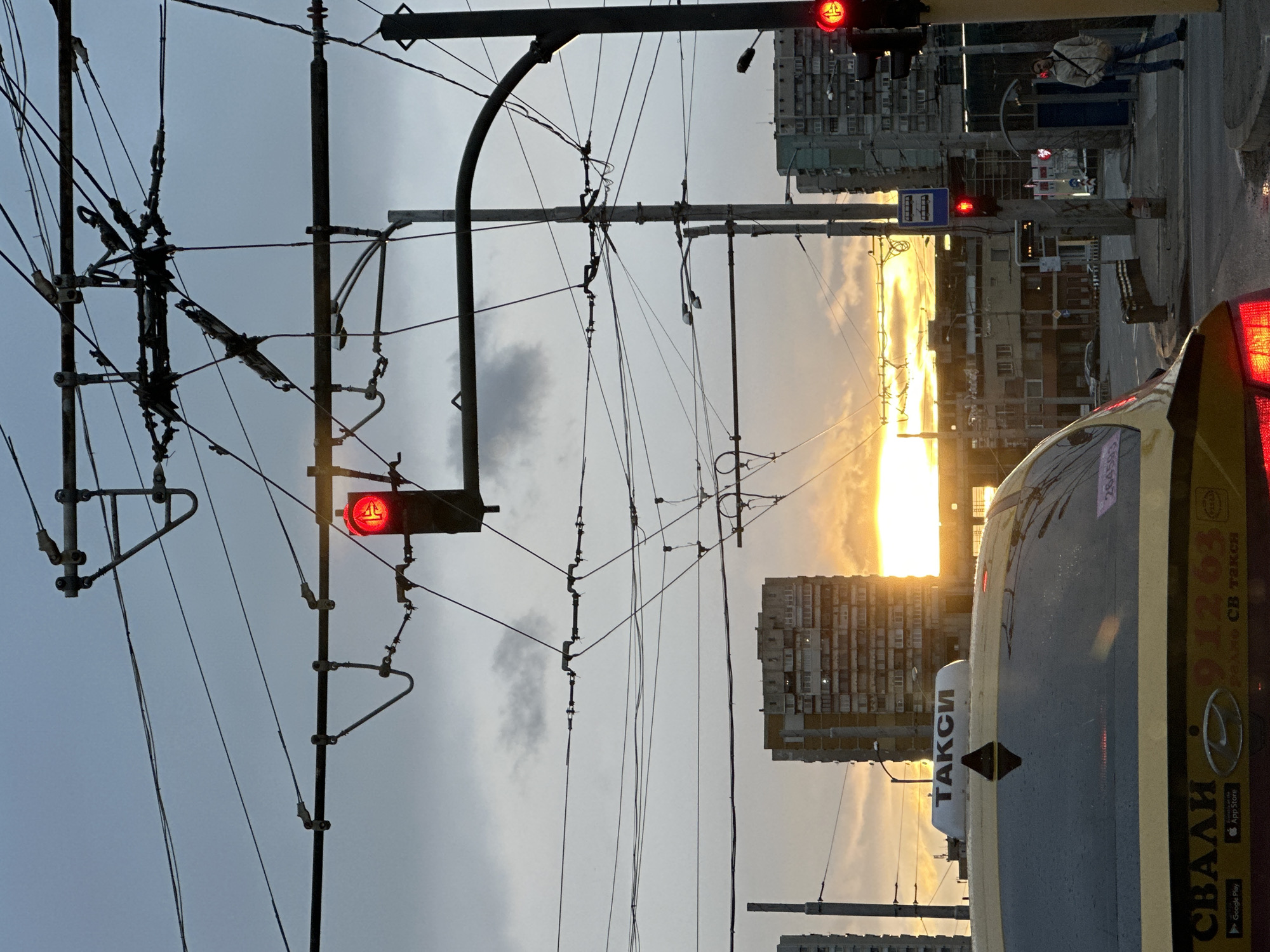 The image captures a street scene in Sofia, Bulgaria at sunset. Overhead, a complex network of tram and trolleybus wires crisscrosses the sky. A red traffic light is visible. Buildings line the street, silhouetted against the bright setting sun. A taxi is parked in the foreground; its side is partially visible. A pedestrian is crossing the street.