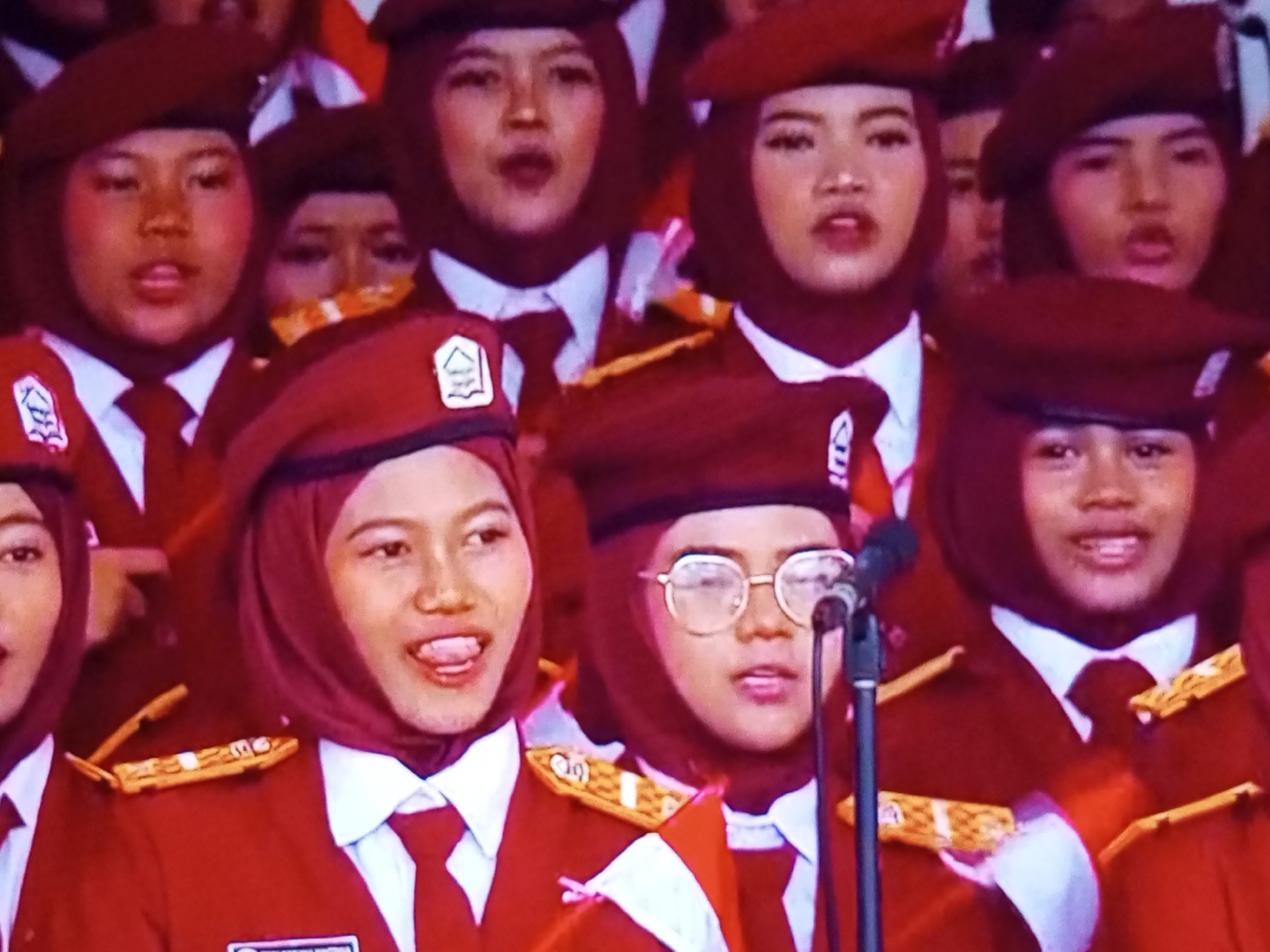 The image captures a group of young women in uniform, likely students, standing together. They are wearing red berets and red blazers with white collared shirts and red ties. Some of the women are wearing hijabs underneath their berets. There are badges or insignia on their blazers, and one woman in the foreground is wearing glasses. The background appears to be a performance or assembly area, with some suggestion of a stage or seating. The lighting is bright, suggesting an indoor or well-lit outdoor setting. One of the individuals in the foreground is sticking her tongue out. The image seems to be a candid or posed shot of the group during an event.