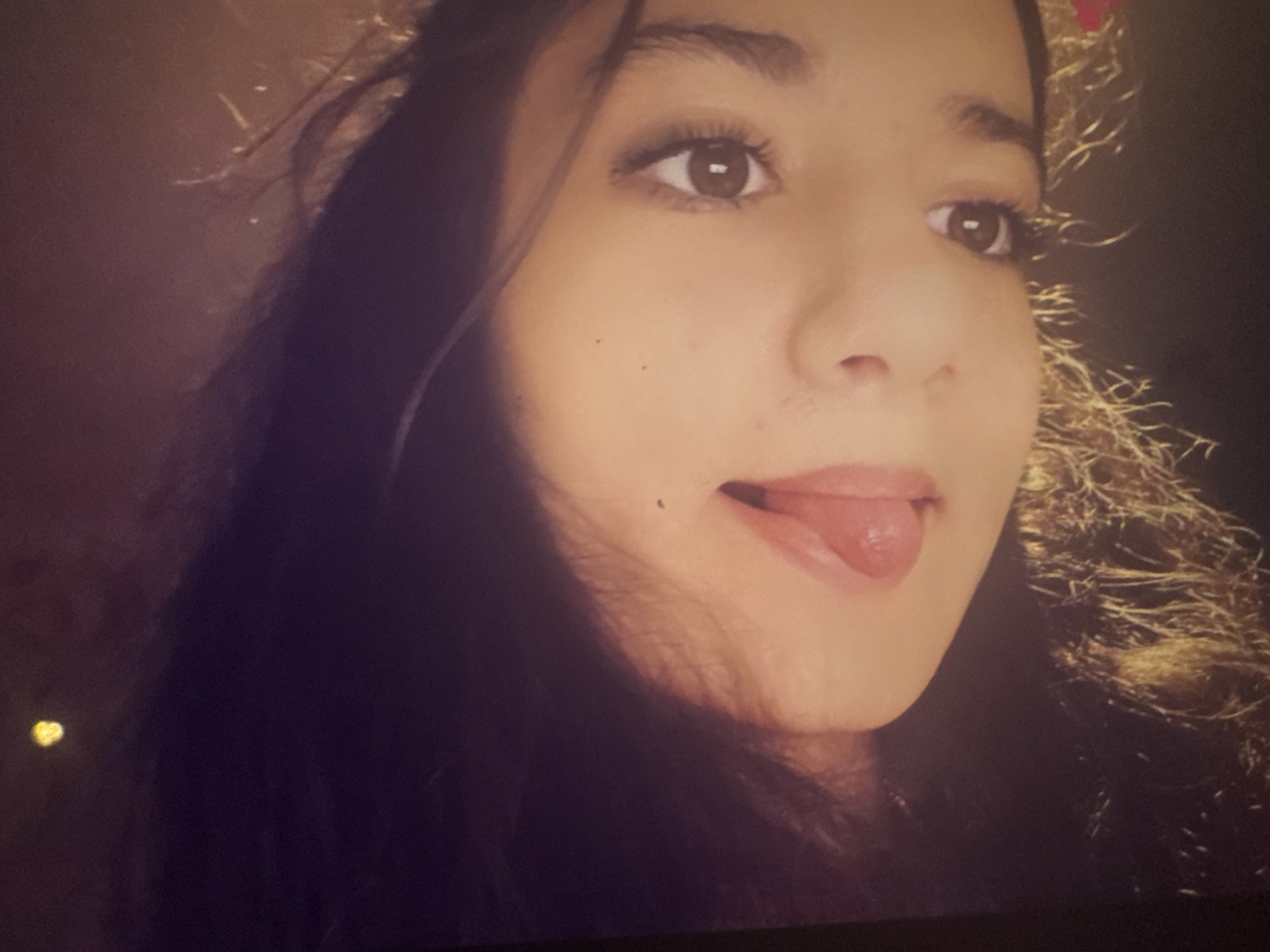 This is a close-up, dimly lit portrait of a young person, likely female, with dark, long hair and a medium skin tone. The subject's face is prominent, and they are looking slightly upwards and to the right, with their tongue playfully sticking out from the corner of their mouth.
The subject has dark eyes, which appear to have mascara or eyeliner. A small beauty mark or mole is visible on their right cheek (viewer's left), beneath the eye. Their hair is dark and falls around their face. On the right side of the image (the subject's left side), some strands of hair appear to have a golden, possibly tinsel-like, accessory intertwined with hints of red, suggesting a festive or decorative element.
The background is dark and largely indistinct, implying a low-light environment, possibly indoors or at night. In the bottom-left corner of the original image, a small, bright yellow, heart-shaped light or graphic is visible. The overall mood conveyed is informal and playful due to the subject's expression. There is no visible text.