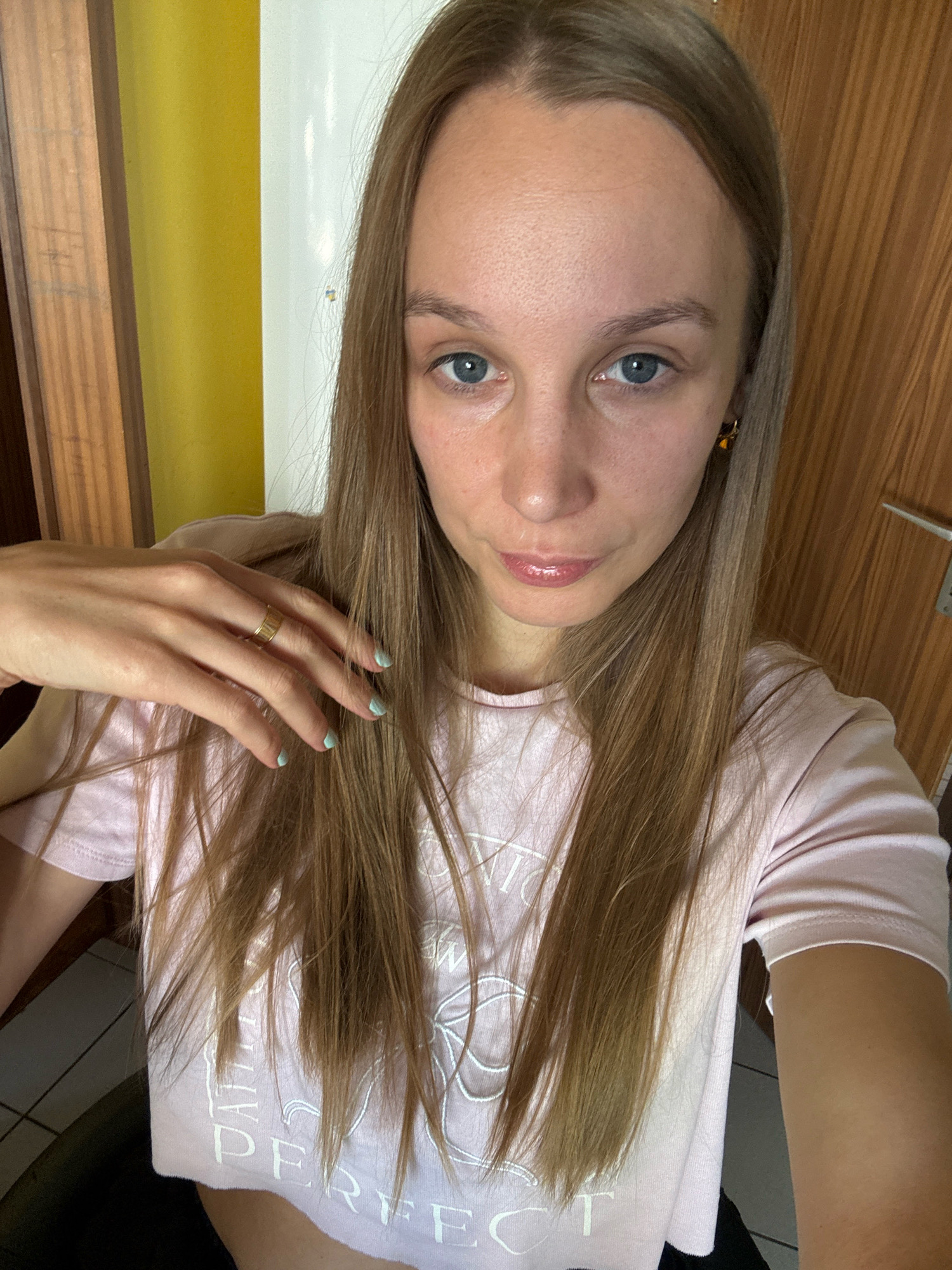The image shows a close-up, selfie-style shot of a young woman with long, light brown hair and blue eyes. She is wearing a light pink crop top with white lettering that appears to say "PERFECT." Her left hand is resting on her chest, and she is wearing a gold ring on her right ring finger and has light blue nail polish on her fingernails. She is also wearing a small gold earring in her right ear. The background is a mix of a wooden door and a yellow wall, with a hint of white in the upper right corner. The overall lighting suggests an indoor setting, possibly a home, given the presence of a door and wall. There is no indication of the image being taken in Überherrn, Germany.