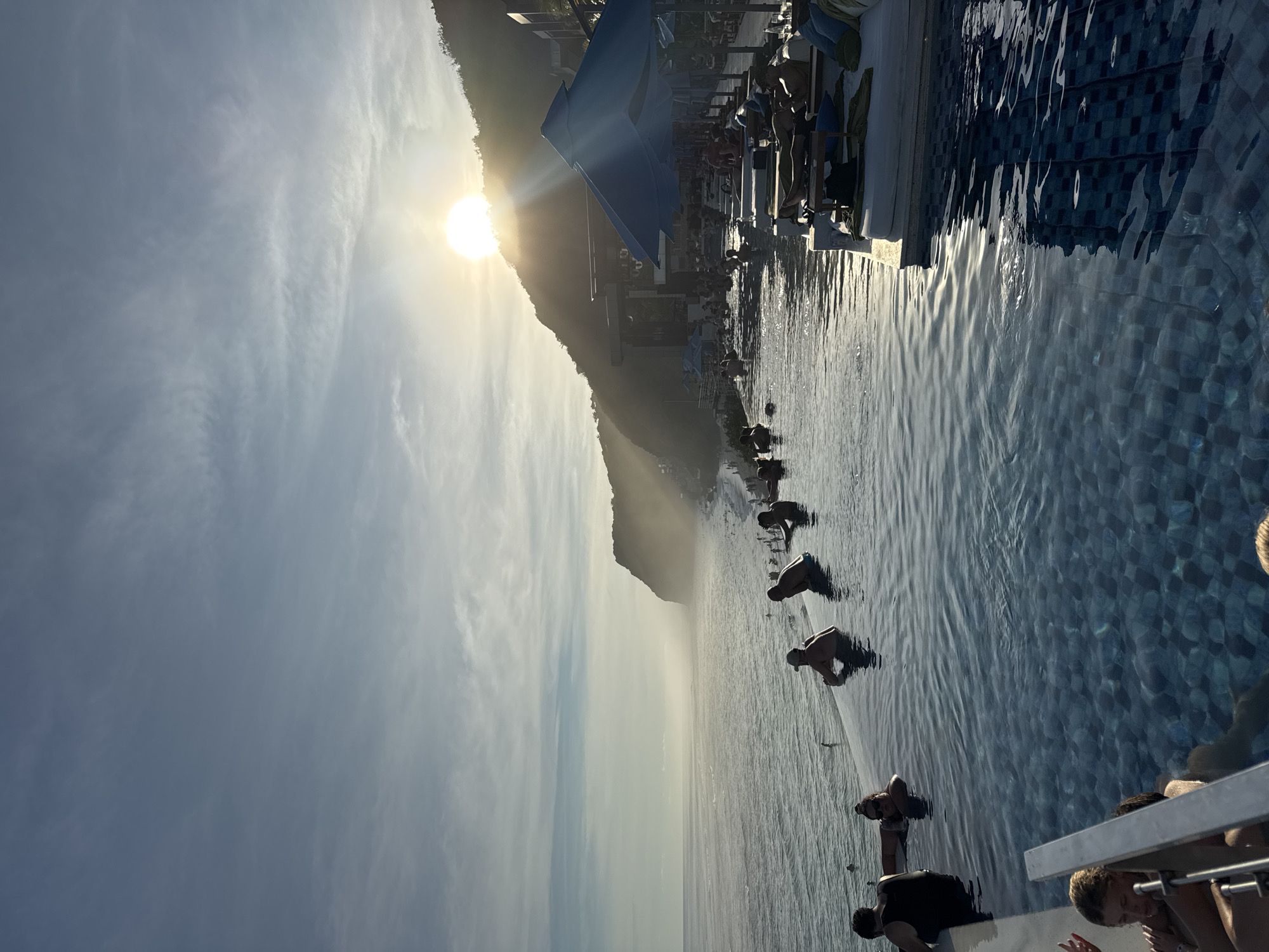The image shows a landscape in Kuta Selatan, Indonesia, featuring an infinity pool overlooking a body of water. The sun shines brightly behind a mountain on the left, casting light across the sky with clouds. Several people are in the pool, some sitting at the edge, others swimming or standing in the water. Lounge chairs and umbrellas are situated on the pool deck. The pool's tiles have a blue mosaic pattern.