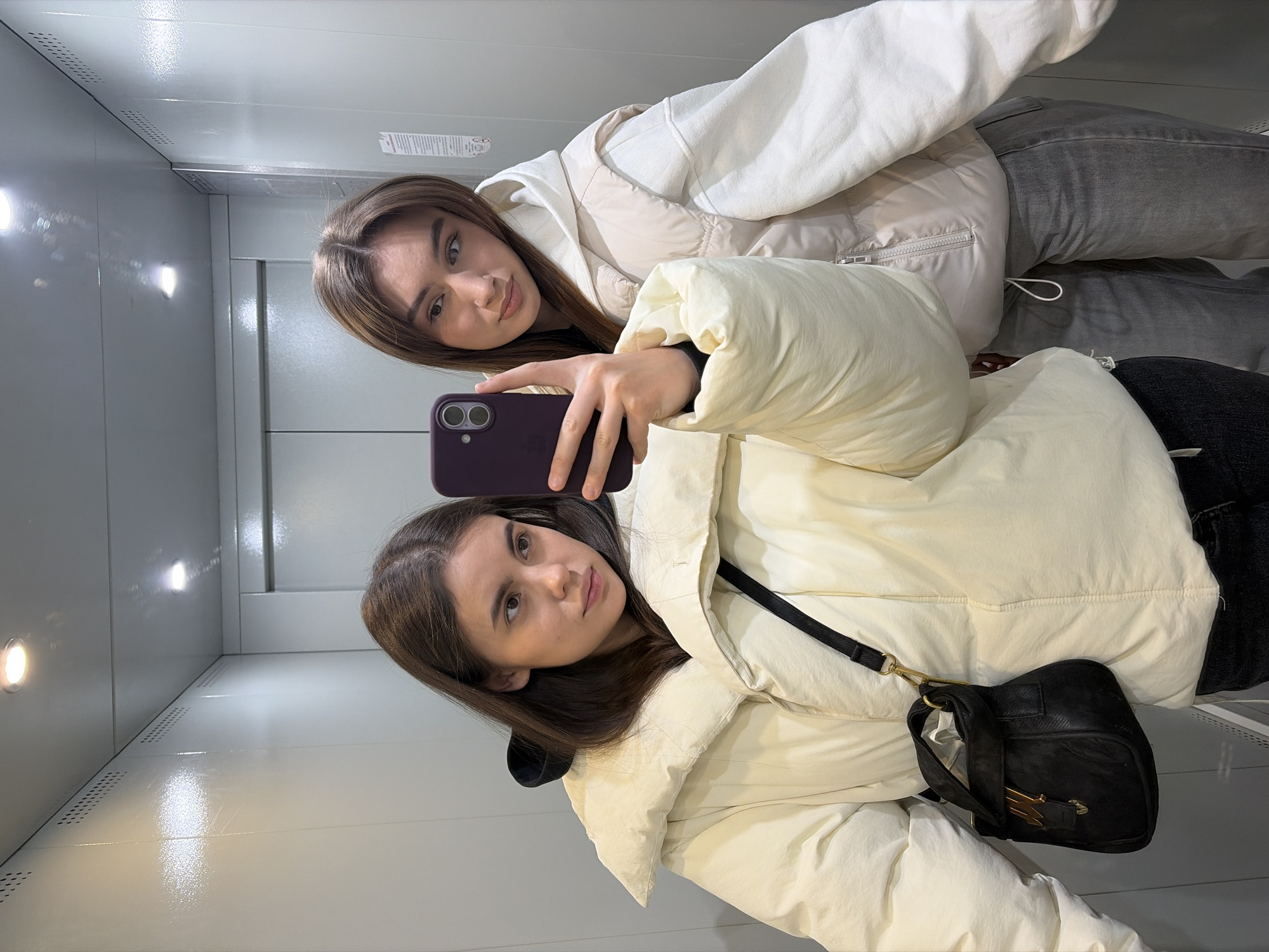 Two young women are captured in a selfie taken inside an elevator, rotated 90 degrees clockwise from a typical upright orientation. The woman in the foreground holds a dark-colored smartphone with a dual-lens camera, extending her right arm to frame the shot. She wears a light cream or off-white puffer jacket and has a black shoulder bag with gold-toned hardware, including a distinctive lightning bolt-shaped clasp. Her expression is neutral, looking directly at the camera.
Behind her, the second woman, also looking at the camera, leans into the frame with a slightly puckered or "pouty" expression. She is dressed in a light beige or cream puffer jacket over what appears to be a white hooded sweatshirt, paired with grey jeans.
The setting is an elevator, characterized by its glossy, light-colored (possibly white or light grey) walls which reflect light. Small, round light fixtures are visible on the walls, casting a warm glow and creating reflections on the polished surfaces. Perforated metal strips or vents are integrated along the edges of the elevator panels, adding to the industrial feel of the space. No specific text is legible on any visible signs or plaques within the elevator. The scene suggests a casual moment, possibly between friends, in Chaikovsky, Russia.