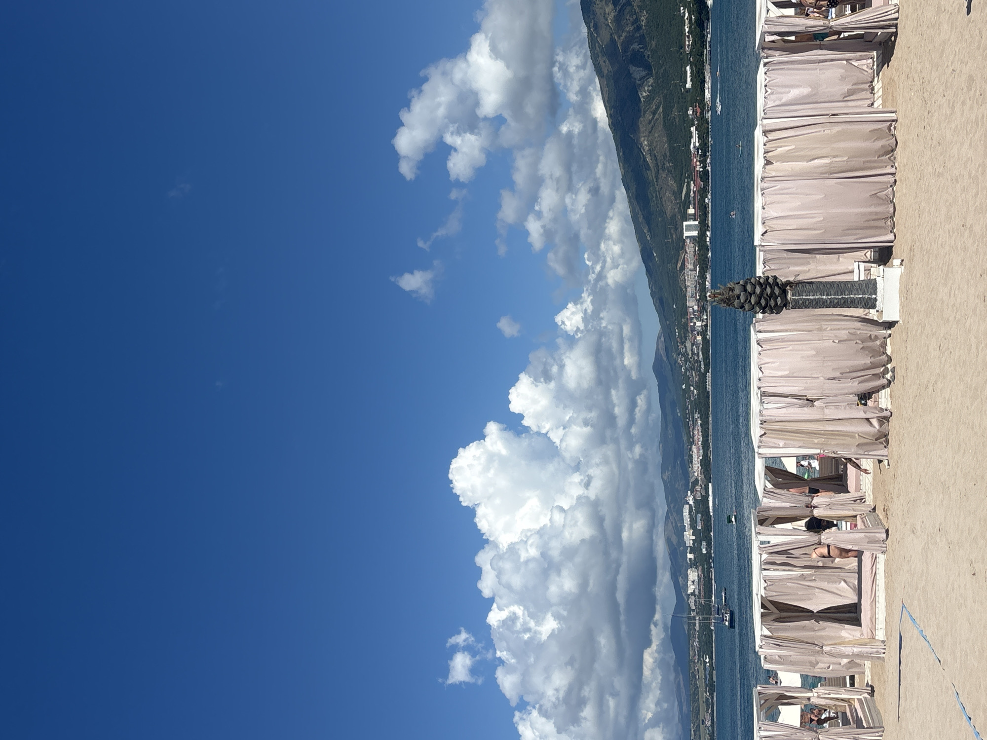 The image depicts a sandy beach with several white cabanas lined up along the shore. The cabanas are furnished with light-colored curtains, and some people are visible inside and around them. In the background, there is a body of water, likely the sea, with several boats and a coastline dotted with buildings and lush green hills. The sky is a vibrant blue with scattered white clouds, and the overall scene suggests a sunny day at a resort or beach town.