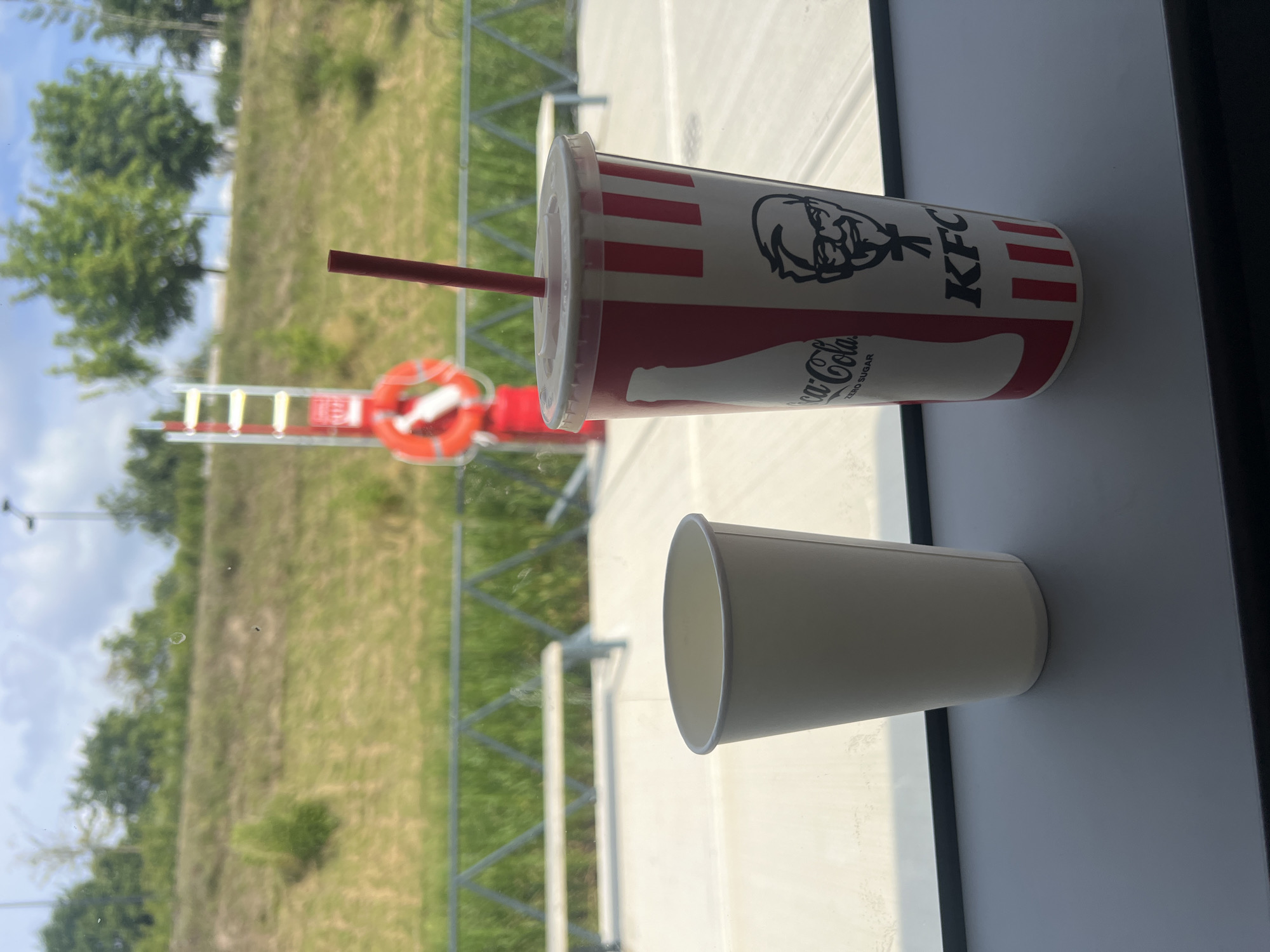 In Vilnius, Lithuania, an indoor scene is presented with two cups on a dark gray ledge. The cup on the left is a plain white paper cup. The cup on the right is a red and white striped paper cup with the KFC logo and a silhouette of Colonel Sanders. A red straw protrudes from the KFC cup. Through a window behind the cups, a grassy hillside is visible, with trees and a bright blue sky with some clouds. A red and white life preserver ring attached to a red pole is also visible on the hillside. In the foreground, a metal railing with a triangular pattern runs horizontally.