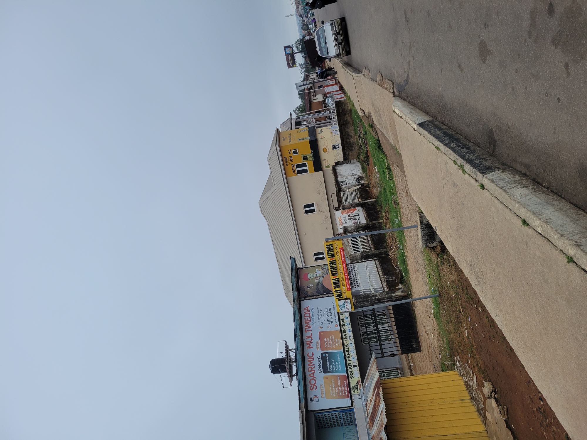 Here is a description of the image:
The image depicts a street scene in an urban area of Unknown, Unknown. The view is angled, showcasing buildings, advertising signs, a sidewalk, and the edge of a road.
On the left side of the frame, there are buildings with signs, notably one for "SOARMIC MULTIMEDIA" advertising prints, branding, and media services. Another sign advertises "SOLAR PANELS INVERTERS," offering sales, installation, and maintenance. The buildings have elements of yellow and beige coloring.
A concrete sidewalk runs along the right side of the image, next to a road. In the distance, cars and people can be seen on the road. The sky is overcast and appears to be a light gray color.