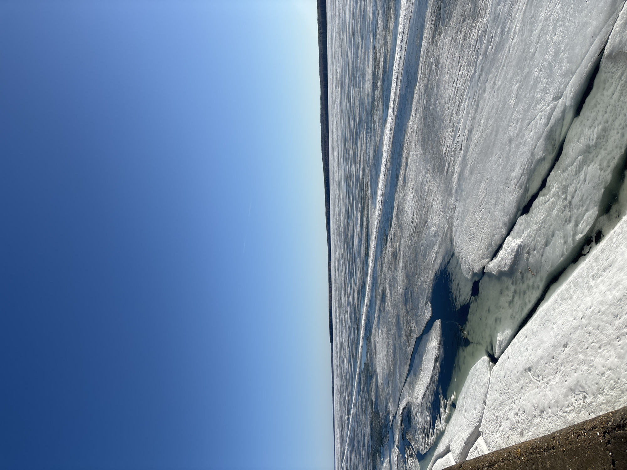 The image shows a frozen body of water under a bright blue sky. Cracks and fissures are visible in the ice, with some areas appearing to have open water underneath. A treeline can be seen on the opposite shore. The concrete edge of a structure is visible in the bottom corner of the image. Based on the location information provided, this is likely the Votkinsk Reservoir near the city of Chaikovsky, Russia, during the winter season.