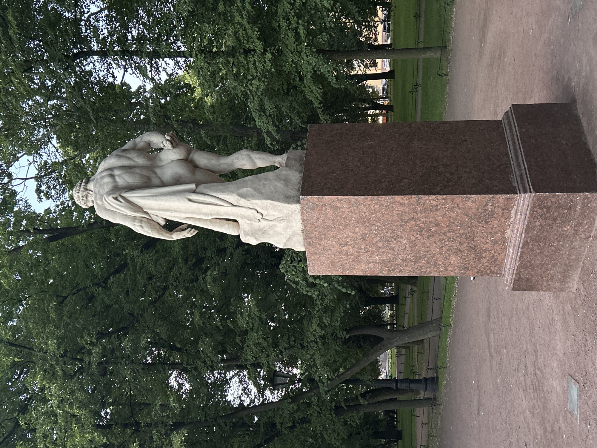 A statue of a muscular male figure, draped in cloth, stands on a granite pedestal in a park. The statue is made of light-colored stone and depicts the figure from behind, with emphasis on the musculature of the back and buttocks. The figure is leaning against a rock-like structure. The pedestal is a rich reddish-brown granite with a layered base. Behind the statue, numerous tall trees with lush green foliage create a dense canopy. A black ornate lamppost is visible to the left of the statue, partially obscured by trees. The ground in the foreground is a reddish-brown dirt path, and a square metal grate is set into the path. In the background, a manicured lawn with pathways and benches is visible, with cars parked along a street beyond the park. The sky is partly visible through the tree canopy, appearing bright and possibly overcast.
