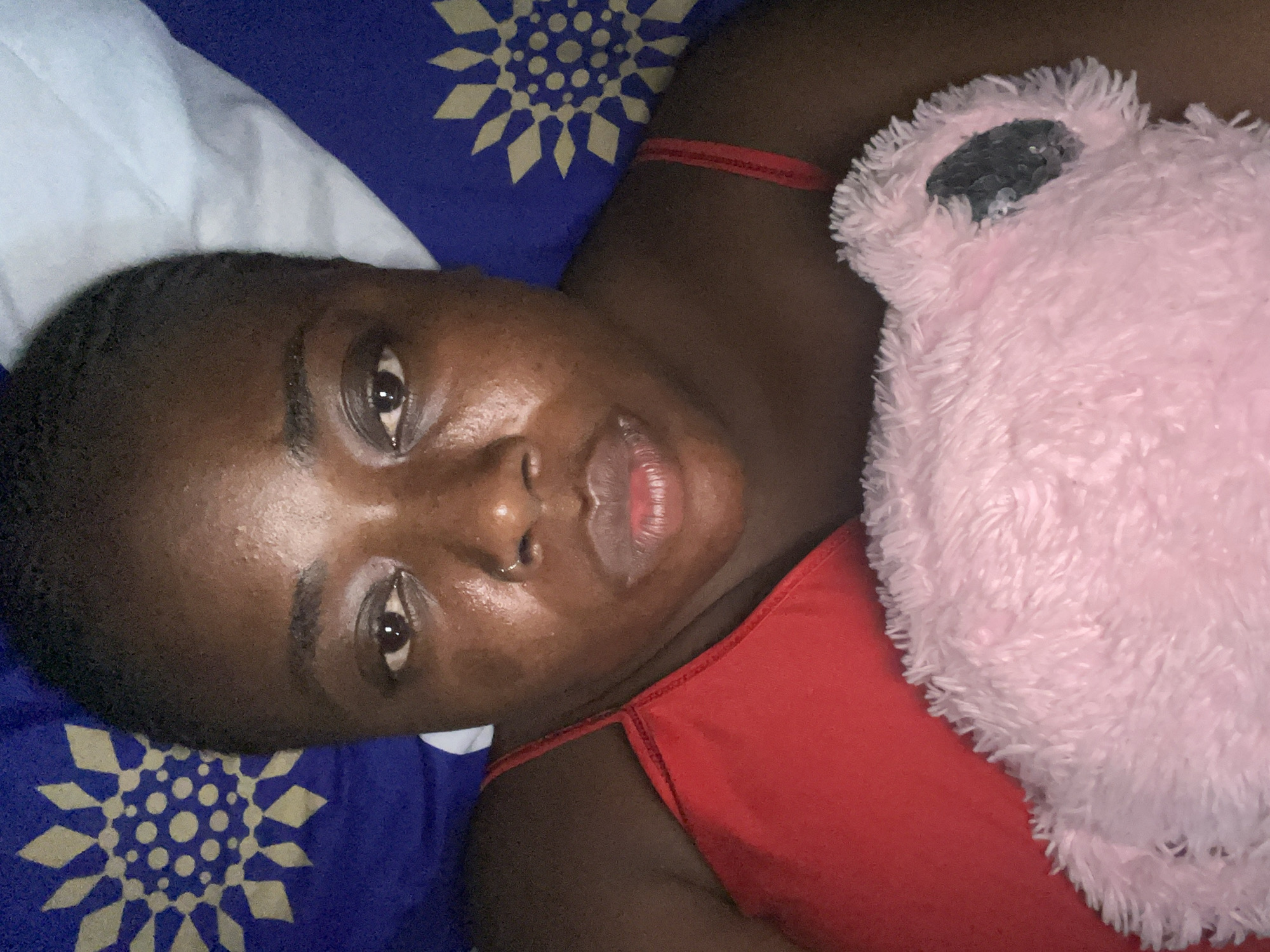 A single individual, an adult female with dark brown skin, is depicted in a close-up, frontal portrait, appearing to be lying down or reclining. She has short, dark hair and wide-set, dark brown eyes, with a visible application of kohl on the lower lash line. A small silver hoop nose ring is present in her left nostril. Her lips are full, slightly parted, and appear moisturized. Her facial skin exhibits a dewy or slightly oily texture with visible pores and some minor areas of hyperpigmentation on her cheeks and forehead. She is wearing a red spaghetti-strap top, visible on her shoulders and chest.
Behind her head to the viewer's right is a light blue pillow. On both the left and right sides of the frame, a dark blue bedsheet or pillowcase with a repeating light gold/yellow circular, star-like geometric pattern is visible. The pattern features a central cluster of small circles surrounded by radiating diamond shapes. She is holding or cuddling a light pink, shaggy-textured plush object, possibly a stuffed animal, which extends across the lower portion of the image. A darker, smooth, possibly sequined patch, likely an eye or ear, is visible on the plush object's right side. The lighting is dim, suggesting an indoor environment. The image contains no visual information to forensically determine a specific city location within Nigeria.