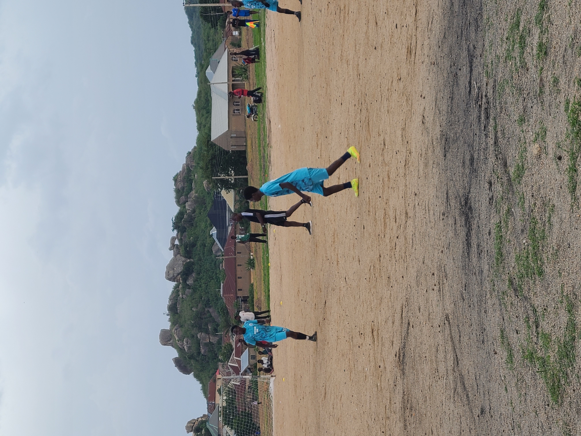 The image shows a group of young men on a dirt field, likely a soccer field, in Jos, Nigeria. The sky is overcast, suggesting a cloudy day. The field is made of sandy soil with patches of sparse green grass. In the background, there are buildings with terracotta roofs and a rocky, tree-covered hillside. Several people are visible on the field, some in light blue soccer uniforms and others in darker clothing. One person in a light blue uniform is in the foreground, appearing to be in motion, possibly practicing soccer drills. Another person in a similar uniform is further back on the field. There are also spectators or other individuals observing the activity. A soccer goal with a net is visible in the lower left corner of the image. The overall scene depicts an outdoor sports activity taking place in a somewhat rugged, natural environment, common in the Jos Plateau region. No visible text is present in the image.
