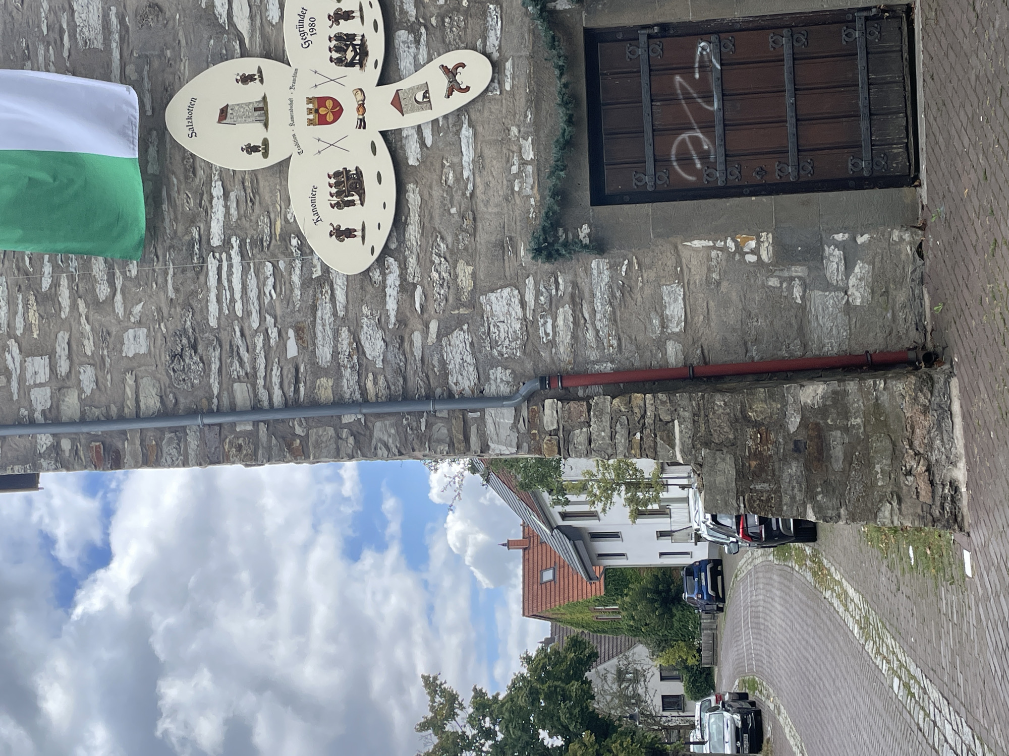 The image shows a street scene in Salzkotten, Germany. On the right side of the image is a stone building with a wooden door. Above the door is a decorative sign shaped like a cloverleaf with illustrations and text. One section of the sign reads "Salzkotten" and depicts a tower. Another section reads "Kanonier" and shows a group of people with a cannon. Another section has the text "Tradition - Kameradschaft - Brauchtum" and a crest. A final section reads "Gegründet 1980" and shows people seated at a table. Above the sign, a green and white flag is attached to the building. The street in front of the building is paved with cobblestones. Cars are parked along the street, and houses with red-tiled roofs are visible in the background. The sky is partly cloudy.