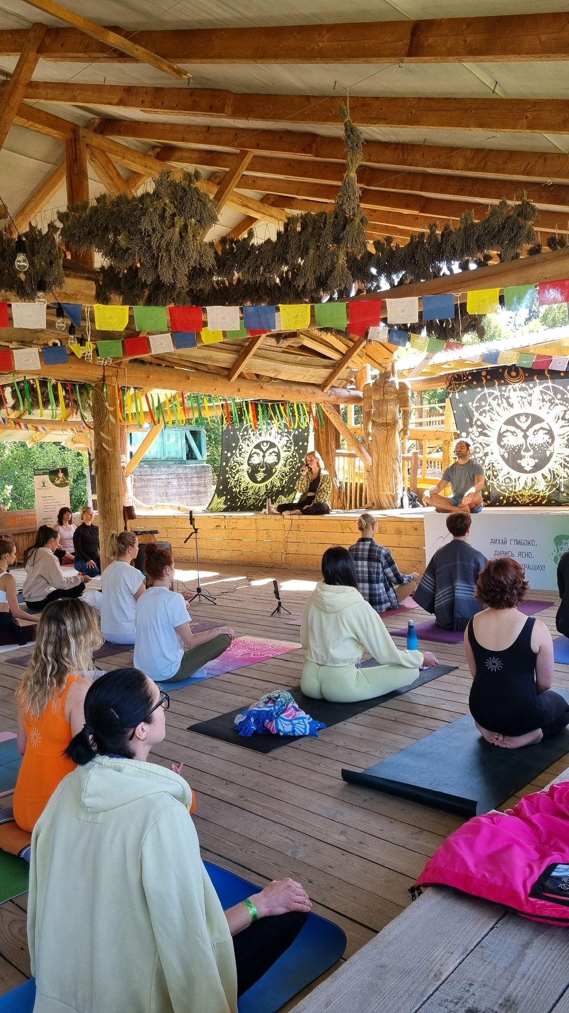 The image depicts an outdoor yoga class taking place under a rustic wooden pavilion in Izky, Ukraine. The pavilion features a thatched roof and is decorated with dried herbs and colorful prayer flags. A stage is set up at the rear of the pavilion, adorned with a black tapestry featuring a sun symbol. Two instructors, a woman and a man, are seated on the stage, addressing the class. Approximately twenty participants are seated on yoga mats on the wooden floor, facing the stage and engaged in meditation or relaxation. The participants are diverse in age and gender, dressed in casual attire suitable for yoga. The environment appears serene and natural, with greenery visible in the background, suggesting a rural or park-like setting.