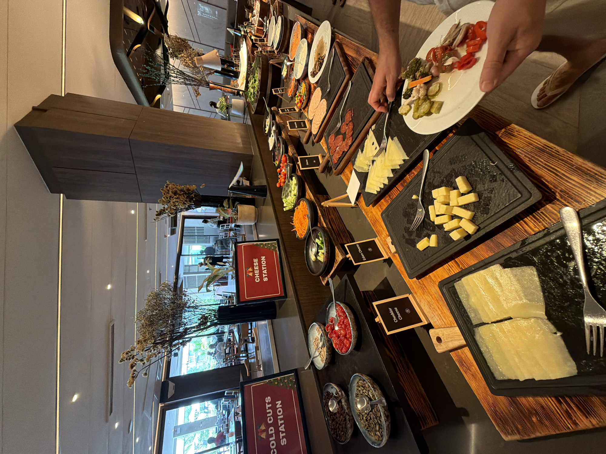 A buffet station is set up in an indoor dining area, likely a restaurant or hotel, in Bang Muang, Thailand. The buffet features various food items, including cold cuts, cheeses, and a salad bar. Several people are present, some seated at tables in the background, while others are interacting with the buffet. A person in the foreground is holding a plate filled with various food items, suggesting they are serving themselves. The lighting appears to be a mix of natural light from windows and artificial lighting from above. The overall scene depicts a typical buffet dining experience.