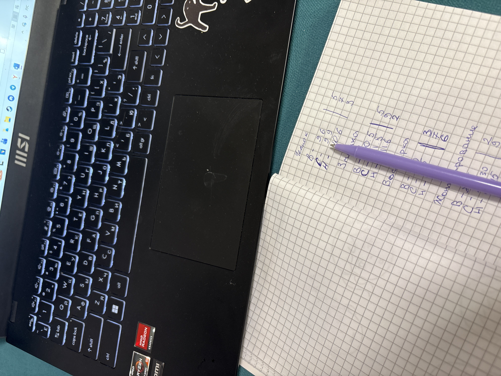 An overhead photograph depicts a black MSI laptop and an open, gridded notebook with a purple pen on a teal-colored surface in Прикамский, Russia.
The MSI laptop, positioned in the upper portion of the frame, features a backlit keyboard with both Cyrillic and Latin characters. Visible keys include 'caps lock', 'shift', 'ctrl', 'alt', 'enter', 'backspace', and a numerical pad. The screen, partially visible at the top, displays a Windows taskbar with various application icons (e.g., browser, folder, Steam, YouTube) and system tray indicators for battery, Wi-Fi, sound, and Russian language input ("РУС"). The laptop's palm rest area displays "AMD RYZEN" and "AMD RADEON GRAPHICS" stickers on the lower left, and an "HDMI" label is partially visible. A black cat silhouette sticker adheres to the lower right corner of the laptop's base. The black touchpad shows minor surface wear.
Below the laptop, an open notebook with a light grey square grid pattern lies on the teal surface. Handwritten notes in dark ink, primarily in Cyrillic, are visible on the right page. A purple pen rests diagonally across the left page, extending onto the right page, obscuring some text. The visible text includes categories and numerical data:
* "Человек" (Person) with associated values "В - 9.6", "С - 9.9", "Н - И0.6", and a column of numbers "5, 7, 3".
* "Кошка" (Cat) with values "В - 0.5", "С - 1.5", "Н - 11.6", and a column of numbers "9, 6, 2".
* "Орка" (Orca, or part of a longer word) with a column of numbers "3, 7, 6".
* Partially visible words such as "3м..." and "...рование" also have associated numerical pairs. Below "...рование", the numbers "30, 15" and "2, 9" are legible.
The surface beneath the objects is uniform in color and texture. No individuals are present in the image.