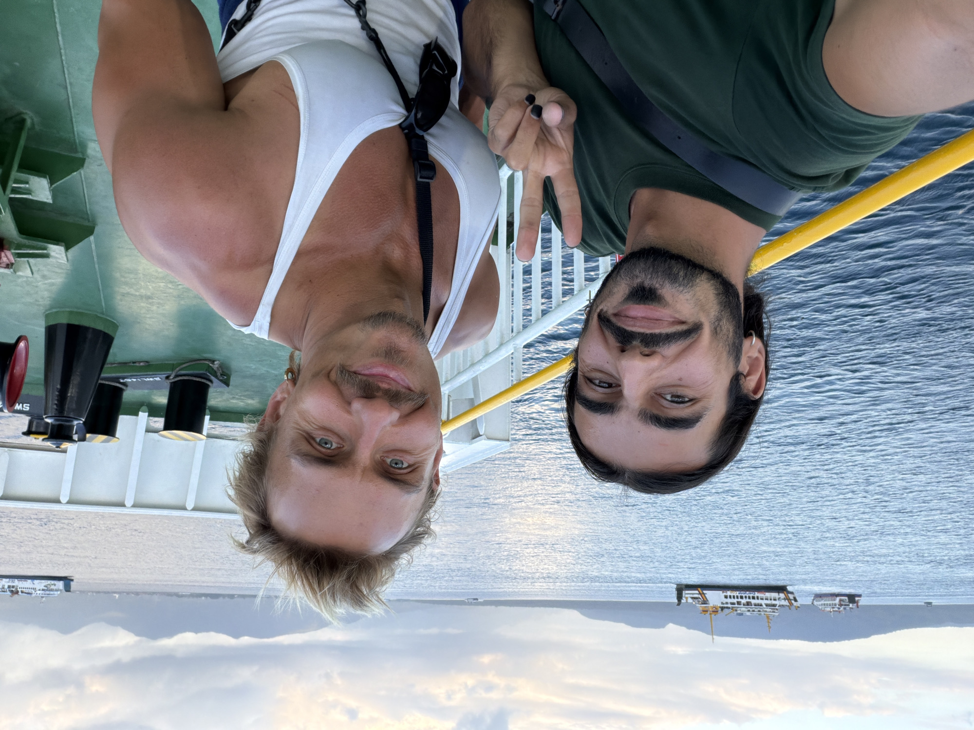 Two men are taking a selfie on the deck of a ferry in the water. The man on the left is wearing a dark green t-shirt and has dark hair and a mustache. He is holding up a peace sign with his left hand. The man on the right is wearing a white tank top and has blonde hair and a mustache. He is smiling. In the background, there are several ferries and islands. The sky is cloudy. The scene is consistent with the port of Ketapang, Indonesia, a major ferry terminal connecting Java to Sumatra and Borneo.