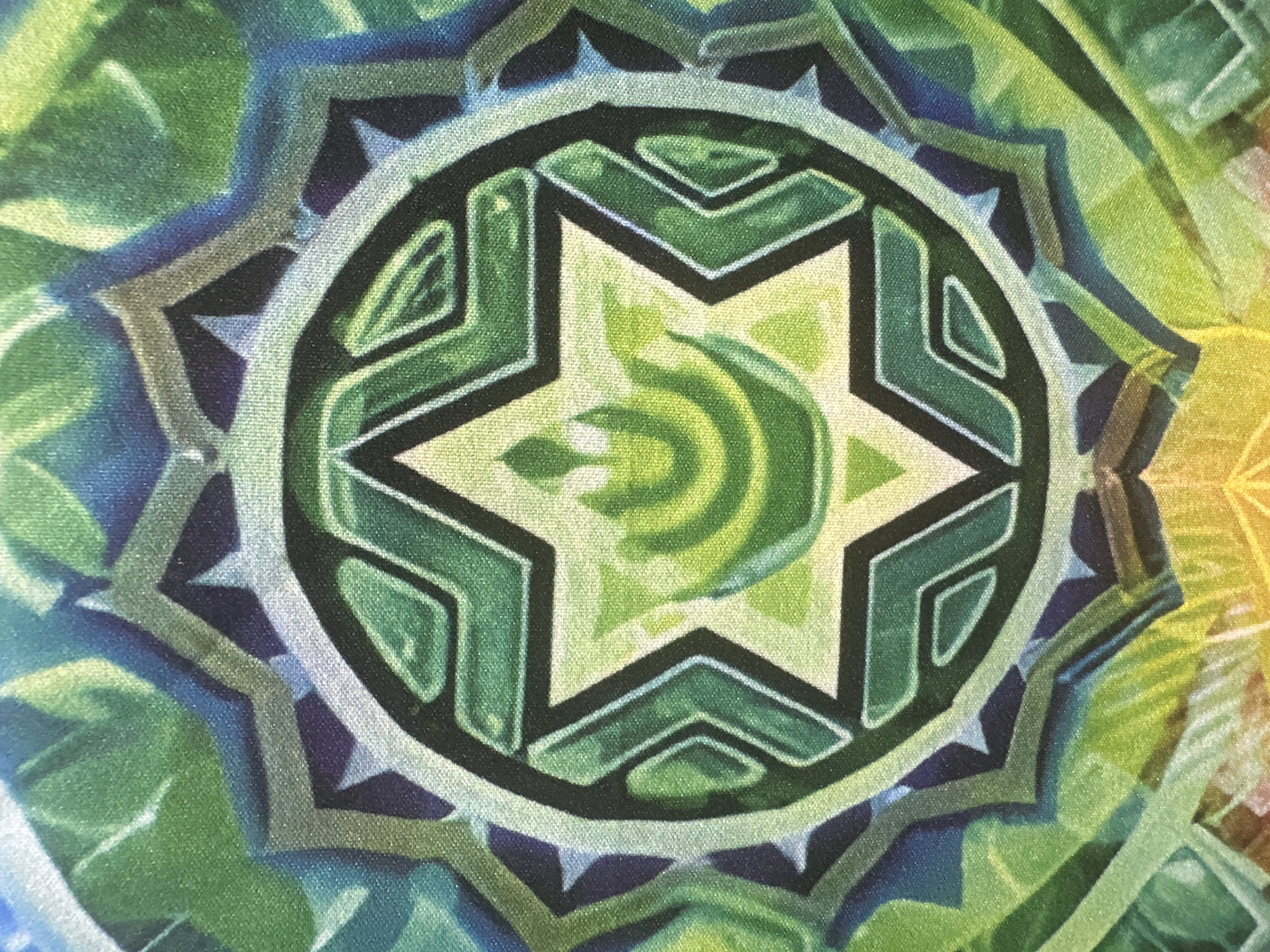 This media file is a close-up, top-down photograph of a fabric tapestry or print. The design is a complex, symmetrical mandala or geometric pattern, rendered in vibrant shades of green, blue, purple, and yellow. The central motif is a six-pointed star with stylized green leaf-like shapes within it. Surrounding the star are concentric layers of geometric shapes, including angular lines and sharp, triangular points. The colors blend and transition smoothly, suggesting a painted or digitally printed design rather than a woven one. The fabric appears to have a slight texture. There is no discernible time of day, weather, location cues, or text visible in the image. The focus is entirely on the abstract, colorful design.