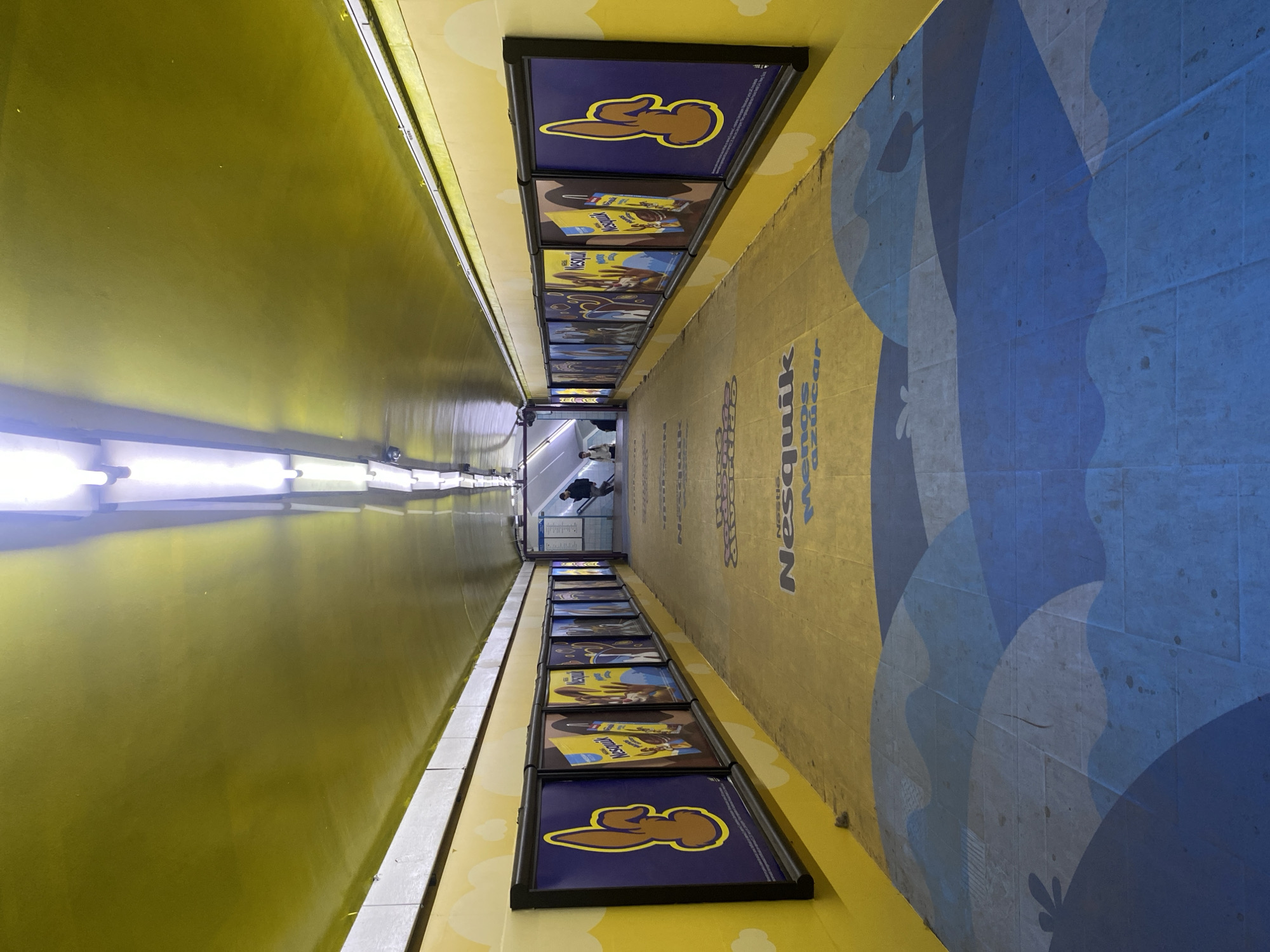 The image shows a brightly lit, narrow hallway in Buenos Aires, Argentina. Yellow walls and ceiling contrast with a blue and tan tiled floor. The floor is decorated with blue wave patterns and Nesquik branding. Framed Nesquik advertisements line both walls. Two people are visible at the far end of the hallway.